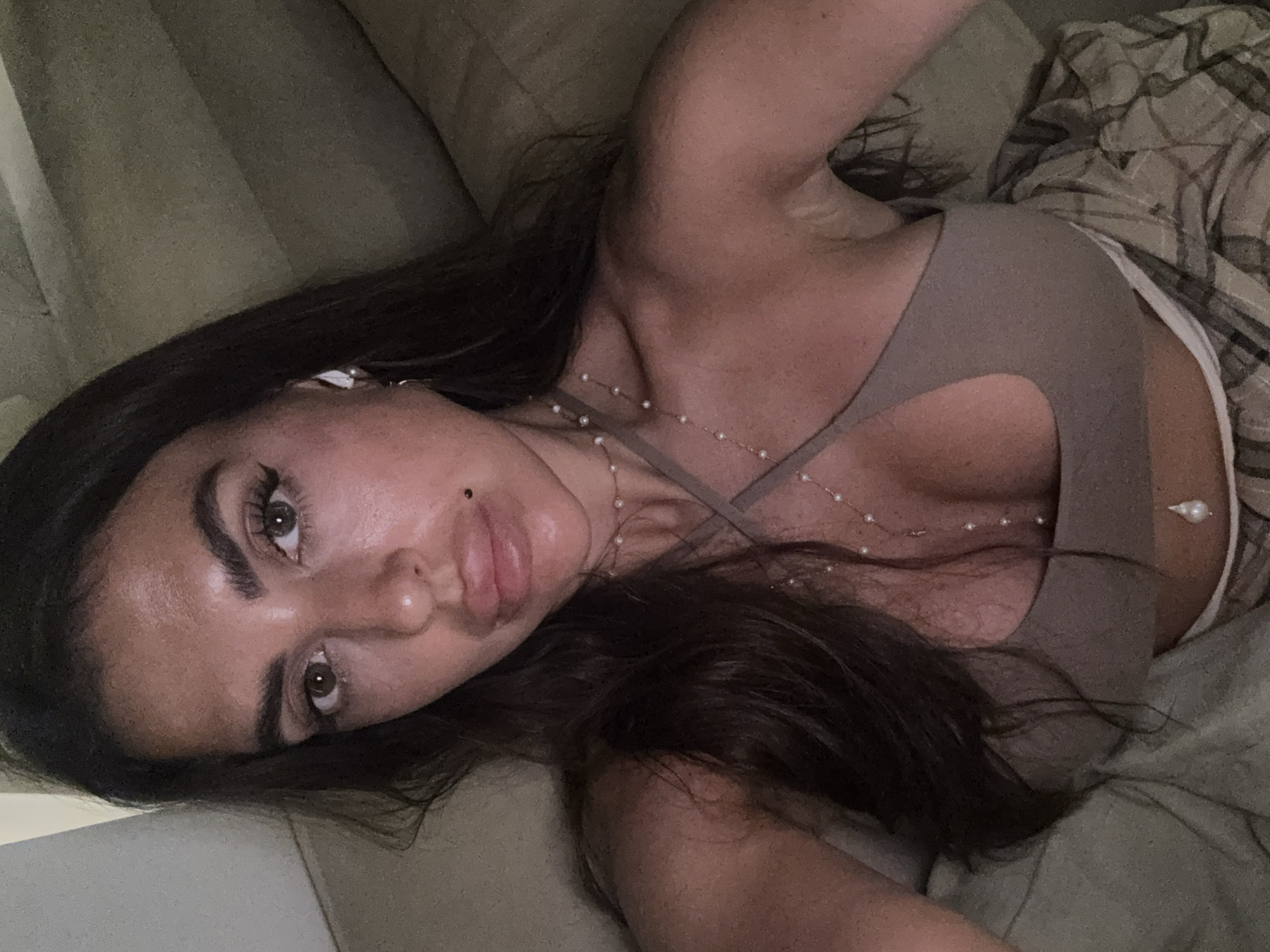 A woman is lying down, possibly on a bed or couch. She has long, dark hair and is wearing a taupe-colored top with a cutout design. She also has a pearl necklace and what appears to be an earbud in one ear. The background includes a pillow and a patterned blanket or clothing. The photo's location is not possible to determine.