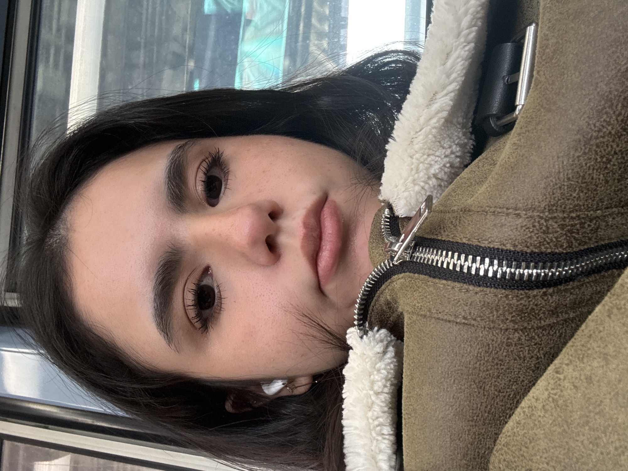A close-up portrait shows a person with dark hair, brown eyes, and fair skin wearing a brown jacket with a white sherpa collar. The zipper of the jacket is visible. The person is likely on a public transportation vehicle, possibly the 'L' train, in Chicago, USA, as suggested by the window frame and urban setting.