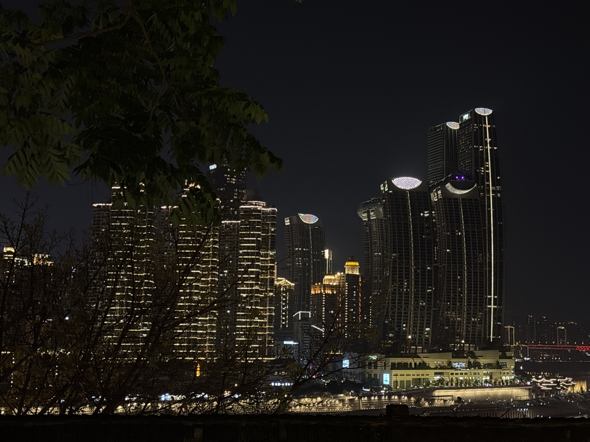 The image shows a night cityscape in Nan'an District, Chongqing, China. Tall, illuminated skyscrapers dominate the skyline, with bright lights creating horizontal and vertical patterns across their facades. The tops of some buildings are adorned with illuminated oval shapes. In the foreground, silhouetted trees and foliage frame the view. Below the buildings, a waterfront area with lights reflecting off the water is visible. A bridge spans the water in the distance. The overall scene depicts an urban environment at night, with a blend of natural and artificial elements.