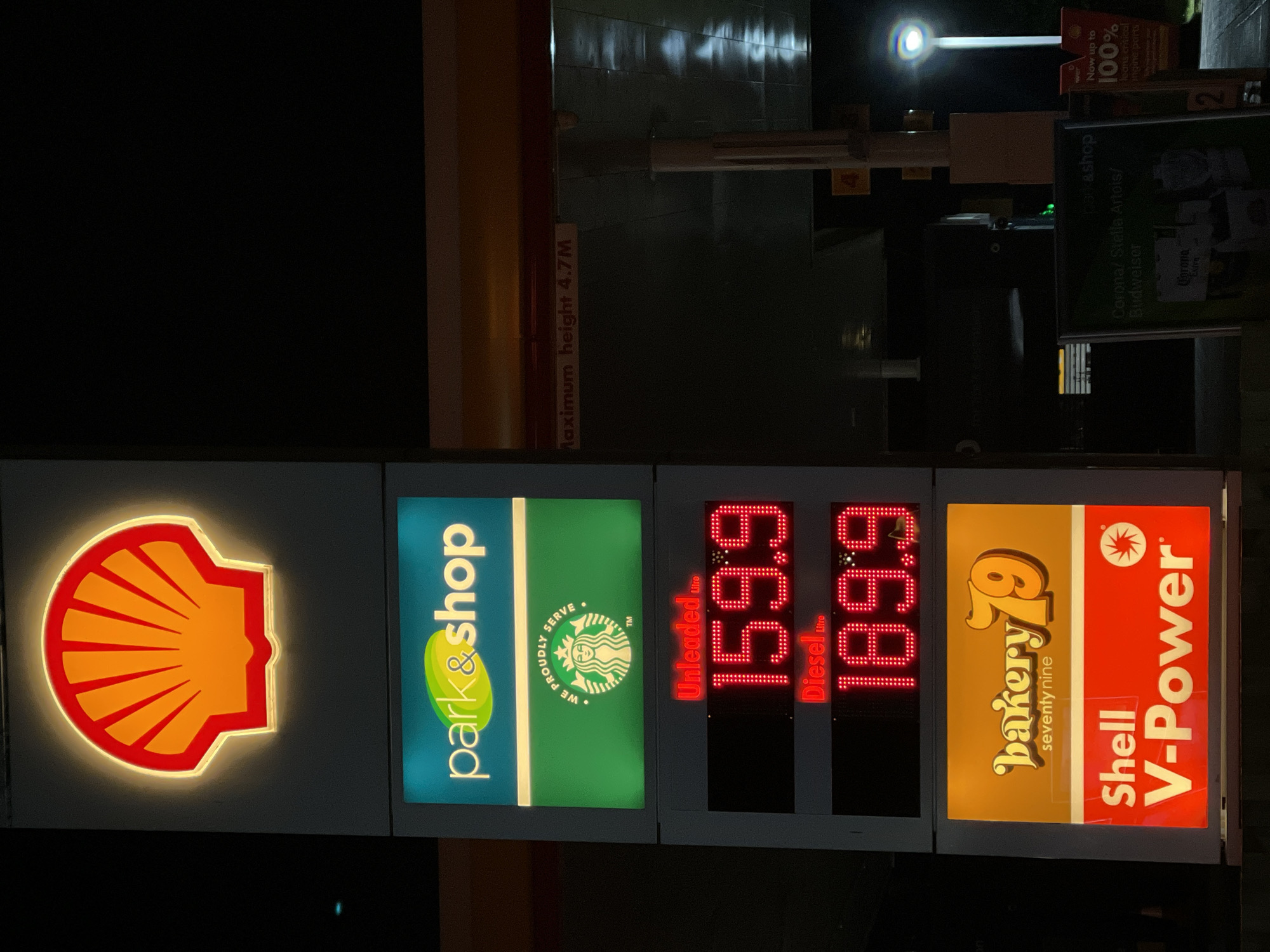 The image shows a Shell gas station in Maidstone, United Kingdom. The gas station's sign displays the Shell logo, a Park & Shop logo with a Starbucks logo, the price of unleaded petrol at 159.9 and diesel at 189.9, and a Bakery 79 logo. Above the sign is a sign with the text "Maximum height 4.7M", and other advertisements with the text "Corona/ Stella Artois/ Budweiser" and "Now up to 100%". There is a single light in the background.
