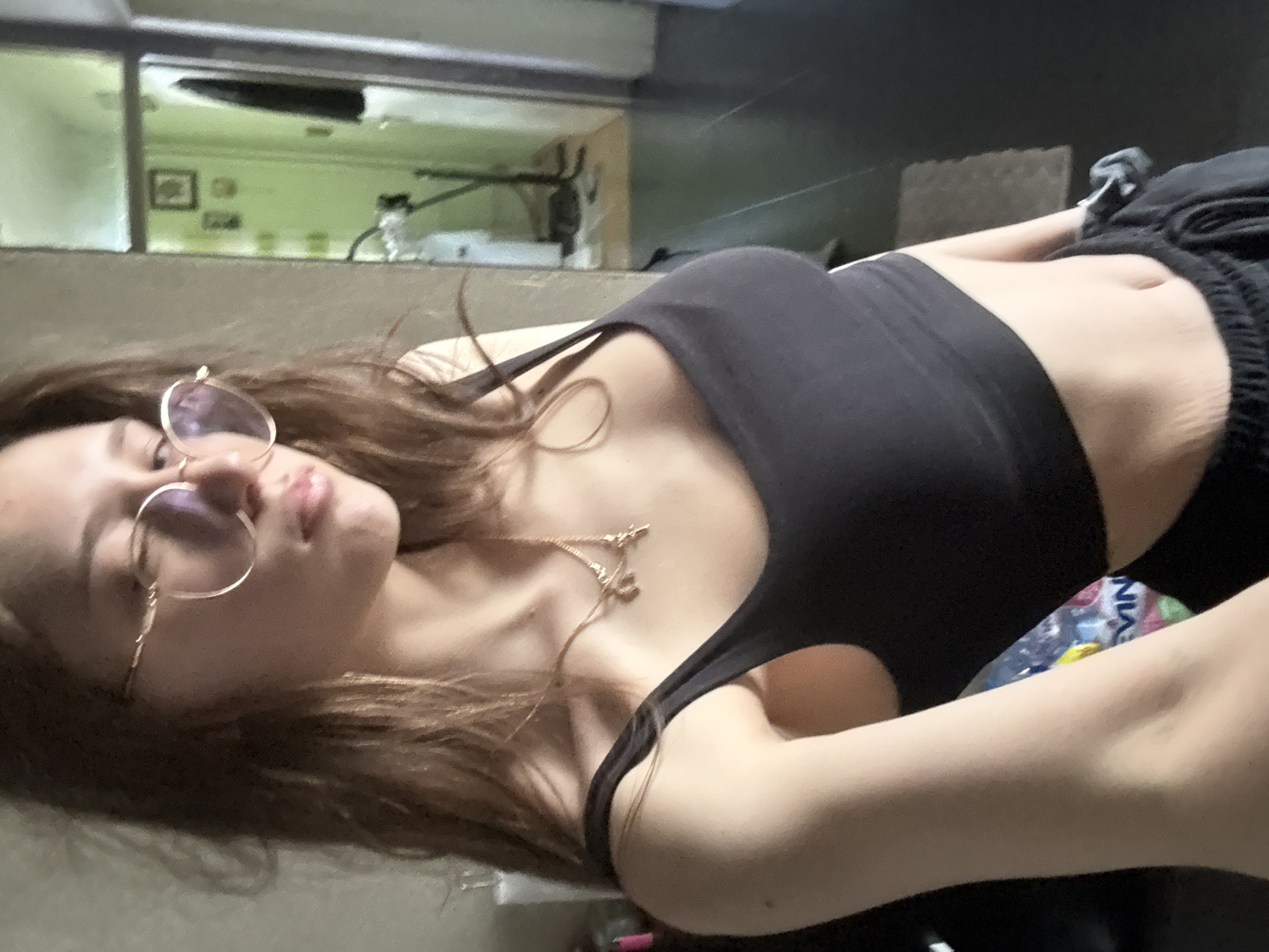 A casual, low-angle indoor portrait features a young woman reclining or lying down. She has long, wavy brown hair, wears round, wire-rimmed glasses, and a delicate gold-colored necklace. Her lips are slightly parted, and her expression is relaxed, looking towards the camera as if taking a selfie. She is dressed in a dark (likely black) tank top or sports bra and dark, casual bottoms, with her midriff exposed.
In the background, behind what appears to be a divider or half-wall, a light green wall with framed pictures or decor is visible. Above this, the background darkens, suggesting an upper wall or ceiling. To the right, near the woman's arm and hip, a plastic bottle with "NIVEA" visible on its label can be seen. The setting conveys a relaxed, everyday atmosphere, possibly within a home in Sofia, Bulgaria. The lighting appears to be artificial, typical of an indoor environment, and the time of day is not discernible.