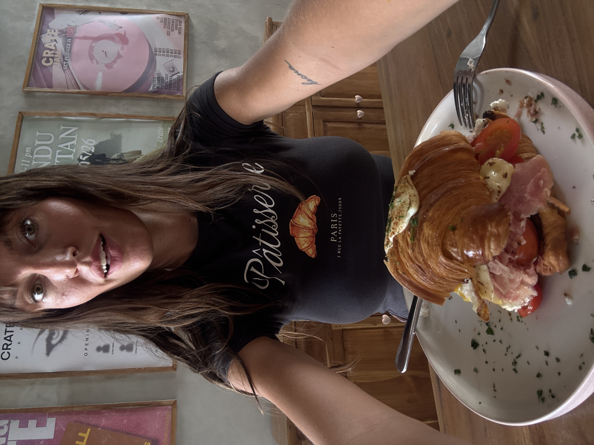 The image shows a woman taking a selfie indoors, likely in a cafe or restaurant in Mengwi, Indonesia. She is wearing a black t-shirt with "Pâtisserie PARIS 3 RUE LA FAYETTE, 75009" printed on it. A plate of food, possibly a croissant with tomatoes and prosciutto, sits on the table next to her. A fork is stabbed into the croissant. There are multiple posters with music-themed designs on the wall behind her. A tattoo is visible on her left arm. The background shows what appears to be wooden furniture.