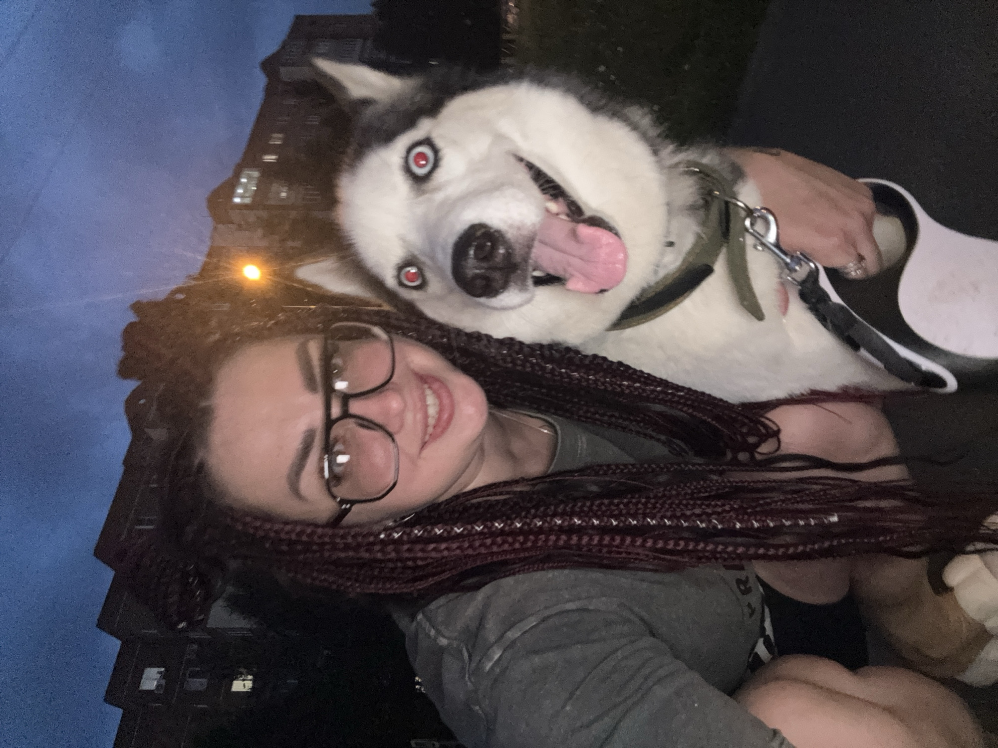 In Tolyatti, Russia, a woman with dark braided hair and glasses smiles for a selfie with her husky. The husky, with striking red-rimmed eyes and a pink tongue hanging out, appears excited. They are outdoors at dusk, with a multi-story apartment building and a street lamp visible in the background, suggesting an urban residential setting. The woman is wearing a grey t-shirt and black shorts, while the dog is wearing a dark green collar with a leash attached to a clip held by the woman. The scene is illuminated by the fading light of dusk and artificial lighting from the streetlamp and building windows.
