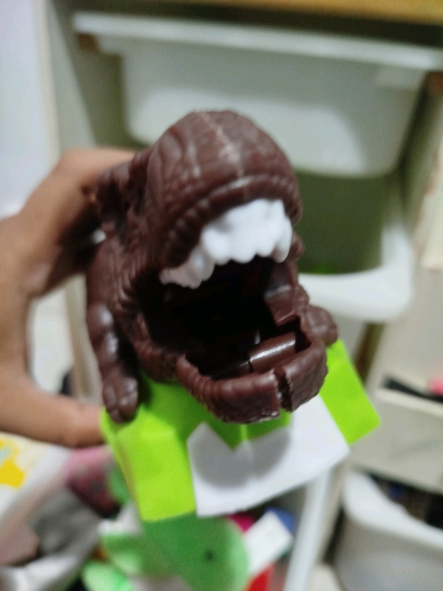 A close-up, slightly blurred image shows a brown, dinosaur-like toy held by a person's hand. The toy has white teeth and an open mouth. It is resting on a bright green object. The background is out of focus, but appears to be an indoor setting with shelves and containers, possibly in a home or storage area. There is no identifiable information to place this image in Mojoagung, Indonesia.