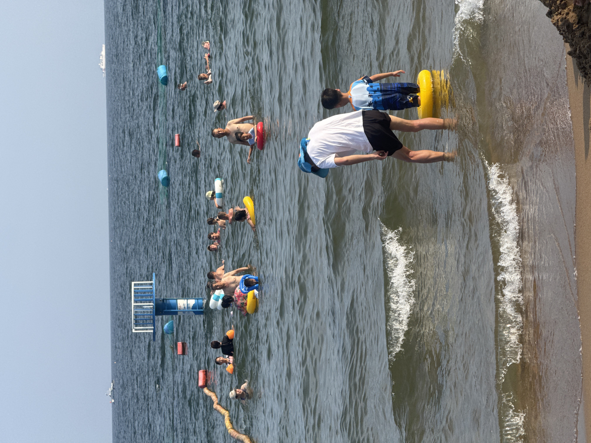 This is a wide-angle, eye-level shot of a crowded beach scene on a sunny day, likely at a public beach in Qinhuangdao, China, given the presence of a lifeguard tower and buoys typical of designated swimming areas. In the foreground, two figures, an adult wearing a white shirt, black shorts, and a blue hat, and a child in a blue patterned swimsuit, stand in shallow water. The child is partially submerged in a bright yellow inner tube. The water is choppy with small waves breaking on the sandy shore visible at the very bottom of the frame.
In the middle ground, a multitude of people are in the water, many with inflatable rings or on floats. A blue and white lifeguard tower with a fenced platform is visible in the water, with a red buoy nearby. A yellow rope barrier stretches across the water from the left side of the frame. Several other inflatable buoys, some blue and some red, are scattered throughout the water.
In the background, the dark blue sea extends to the horizon, meeting a pale blue sky. A white ship is visible in the far distance on the right, and another, smaller boat or marker is seen to the left of the lifeguard tower. The overall atmosphere is one of recreation and summer enjoyment, with numerous individuals swimming and playing in the sea.