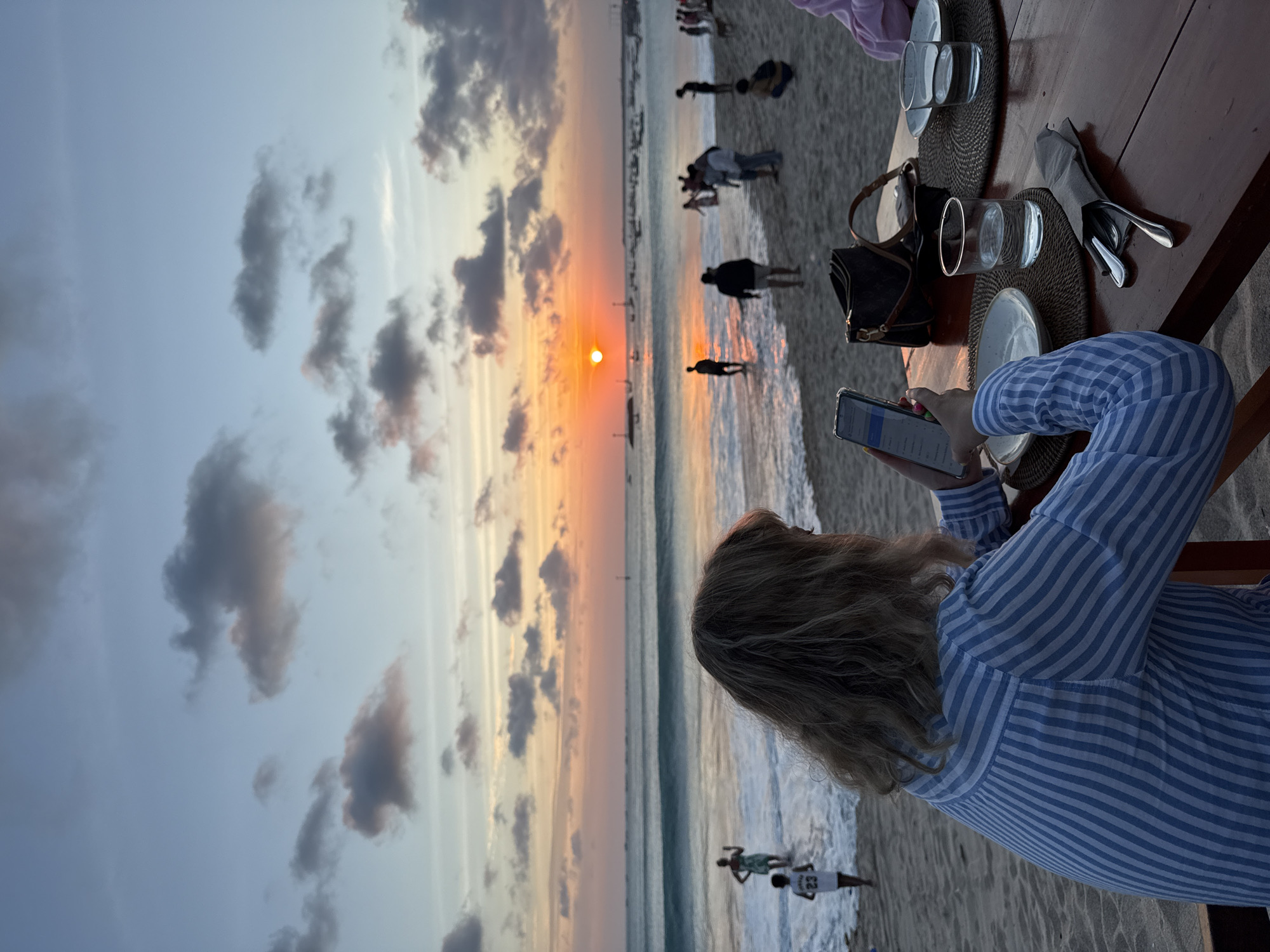 A young woman with blonde hair, wearing a blue and white striped shirt, sits at a wooden table on a sandy beach in Kuta, Indonesia. She is looking at her phone, which is held in her right hand. In front of her are two plates, two glasses of water, and a set of cutlery wrapped in a grey napkin. A dark brown handbag rests on the table beside her.
The beach scene unfolds behind her, with the ocean waves gently rolling onto the shore. Several people are visible in the distance, some wading in the water and others walking along the beach. The sun is setting over the horizon, casting a warm orange and pink glow across the sky and reflecting on the water. The sky is dotted with scattered clouds, some tinged with the sunset colors. The overall atmosphere is serene and picturesque, capturing a moment of quiet contemplation amidst a beautiful natural setting.