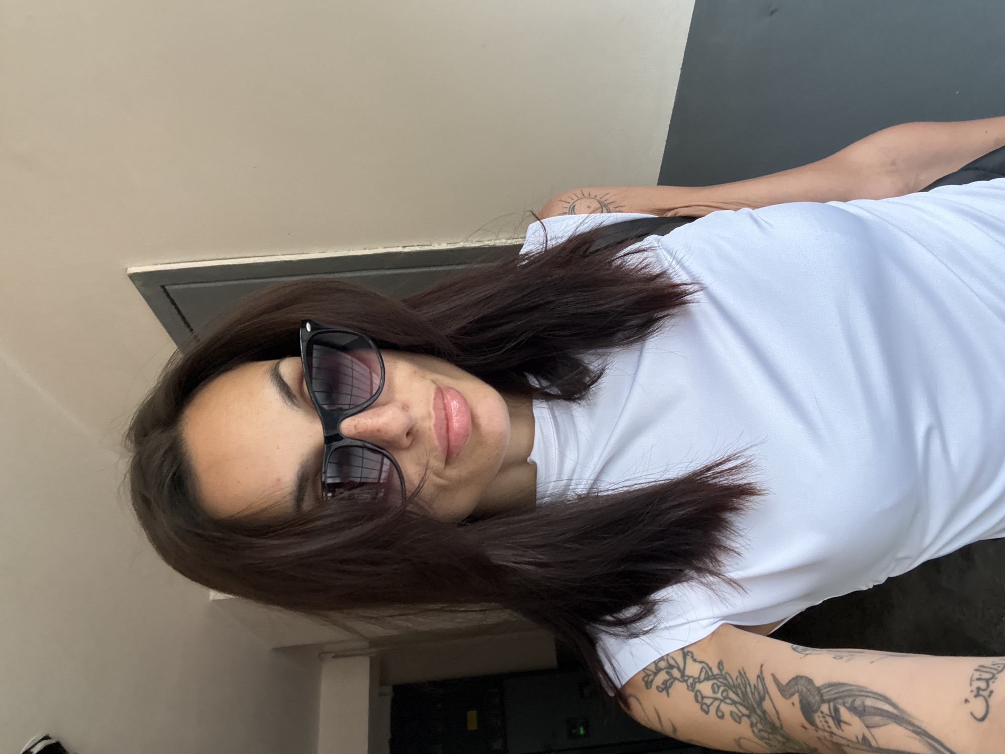 A woman with dark hair wearing sunglasses and a white t-shirt is positioned in front of a wall with a dark gray panel. The woman has tattoos on her arms. The location cannot be determined from the image.