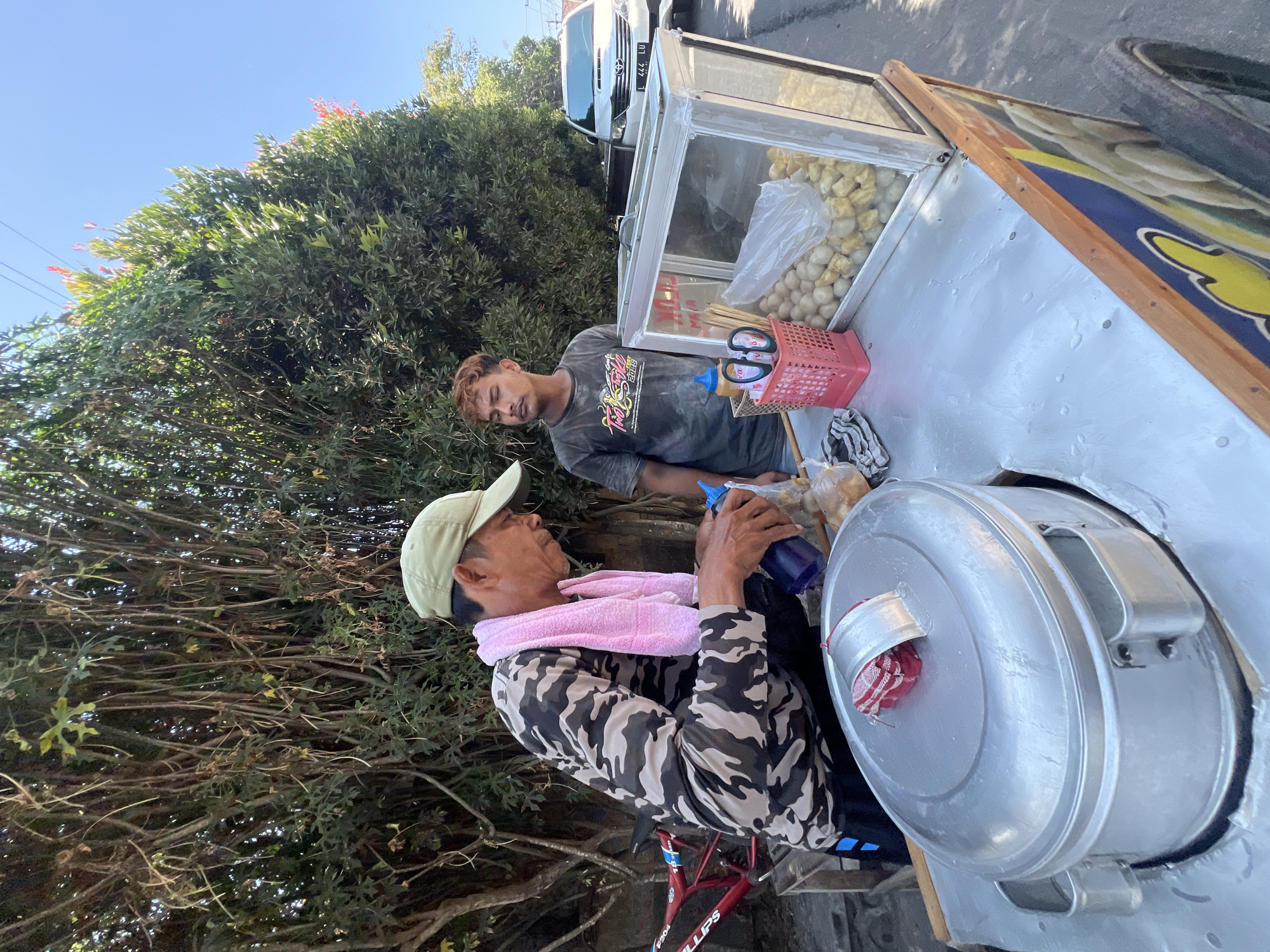 The image depicts an outdoor scene on a sunny day, likely in the city of Denpasar, Indonesia, given the presence of a street vendor and local flora. Two men are positioned around a food cart. The man in the foreground, wearing a camouflage long-sleeved shirt, a light-colored cap, and a pink towel around his neck, is actively preparing food. He is holding a blue bottle, possibly a sauce dispenser, and a plastic bag containing small, round food items. A large, lidded pot is prominently displayed on the cart, suggesting it's used for steaming or cooking.
Next to him stands another man, younger, with styled brown hair, wearing a dark t-shirt. He is observing the activity. The food cart itself is a mobile unit with a display case filled with food items, including what appear to be meatballs or similar snacks. A pink basket containing cutlery and chopsticks sits on the counter. In the background, lush green trees and foliage dominate the scene, with a white car and a red bicycle also visible. The overall impression is that of a typical street food vendor operation in a tropical urban environment.