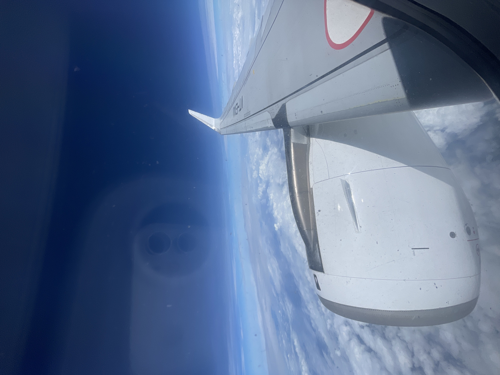 The image shows a view from an airplane window, looking out at the sky and clouds. An airplane wing and engine are visible in the foreground, suggesting the image was taken during flight. The sky is a deep blue, with a layer of white clouds below. There are some smudges and reflections on the window. There is no indication of Neemuch, India, or any urban environment. The image is a generic aerial view from an aircraft.