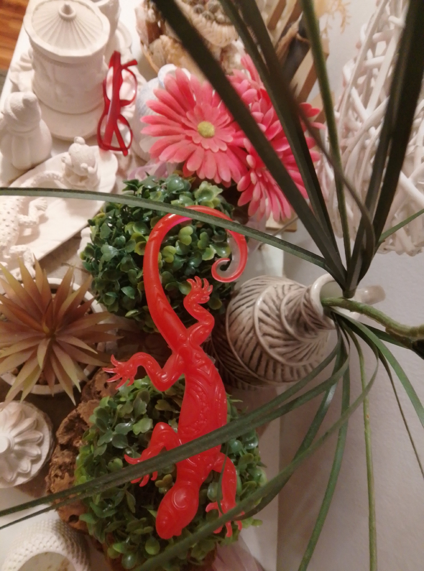 The image depicts a close-up view of a decorative display, likely a tabletop arrangement. The foreground is dominated by a bright red plastic lizard, positioned on top of a clump of artificial green foliage. The lizard is detailed with sculpted limbs and a coiled tail. Behind the lizard, a beige ceramic vase with a textured, twine-like pattern is visible, containing a green plant stem. Further back, pink artificial flowers with yellow centers are clustered, along with a pair of red-framed glasses resting near a white ceramic pot. Other decorative objects, including white ceramic figures and a woven basket with what appears to be dried natural materials, are partially visible in the background. The overall impression is one of a curated still life, with a focus on textures and vibrant colors contrasted with neutral tones. There is no information available regarding the location of this scene in the city of Wedel, Germany.