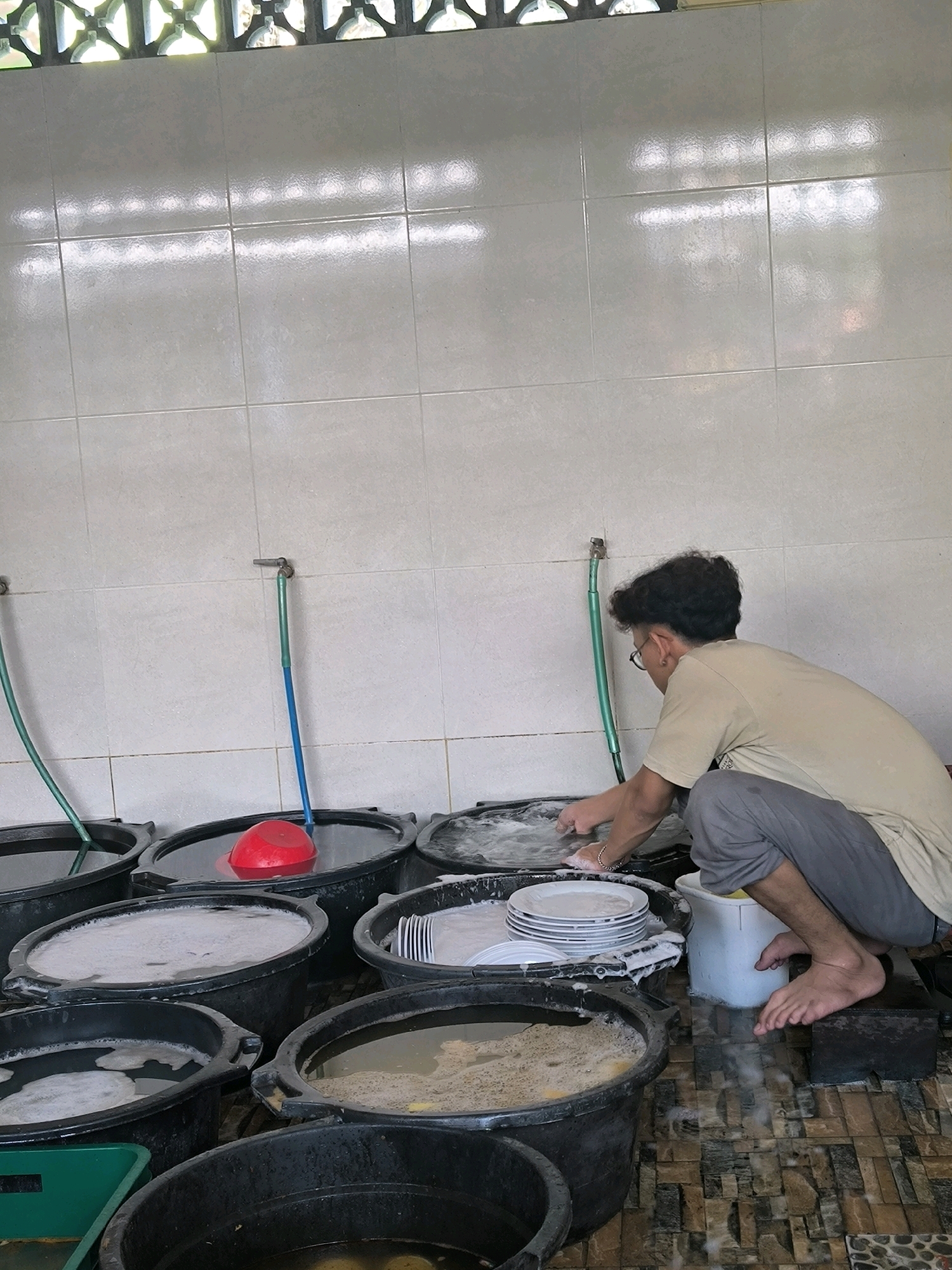 A young man wearing glasses, a light-colored t-shirt, and grey pants is washing dishes in a row of large black plastic tubs filled with soapy water. He is crouching down, with his right hand submerged in the water and his left hand resting on the edge of a tub. A stack of white plates is visible in one of the tubs. The tubs are lined up against a tiled wall, with faucets and green hoses above them. The floor is tiled with a mosaic pattern. The scene appears to be an outdoor or semi-outdoor dishwashing area.