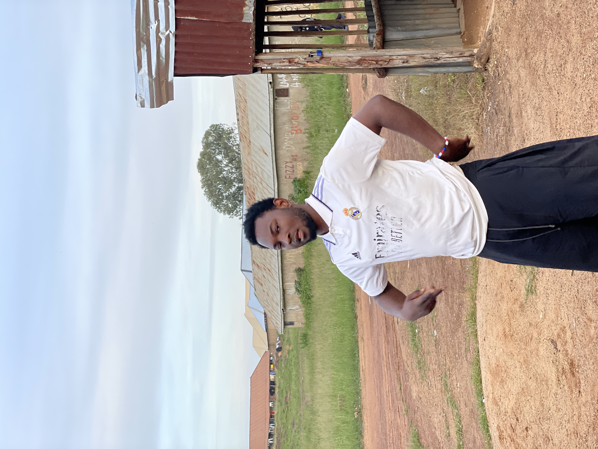 A young Black man stands in a dirt area with sparse grass. He is wearing a white polo shirt with purple accents and black pants. His right arm is bent at the elbow, with his hand near his waist. He has a multi-colored bracelet on his left wrist. Behind him are several buildings with corrugated metal roofs, some appearing weathered. The sky is overcast and light blue. The environment appears to be outdoors in a rural or semi-urban area. The location is the city of Durumi, Nigeria.