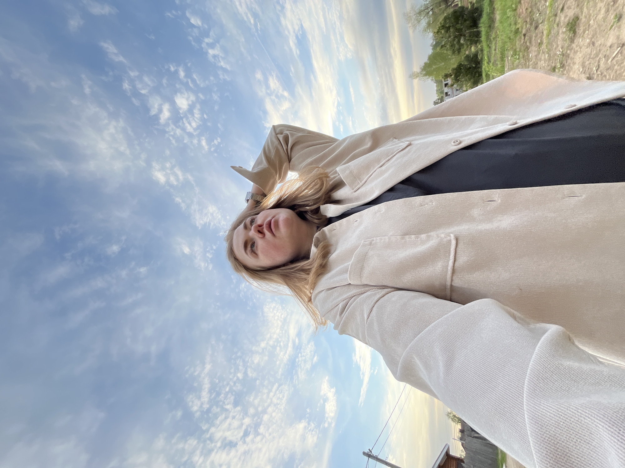 A woman stands outdoors beneath a sky filled with clouds, possibly in the city of Chaikovsky, Russia, though specific landmarks are absent in the image to confirm this. She is wearing a beige corduroy jacket over a black top. The photo is taken from a low angle, pointing upwards towards her and the sky. A wristwatch is visible on her left wrist. The background includes green grass and trees. Utility poles and rooftops are visible in the far distance.