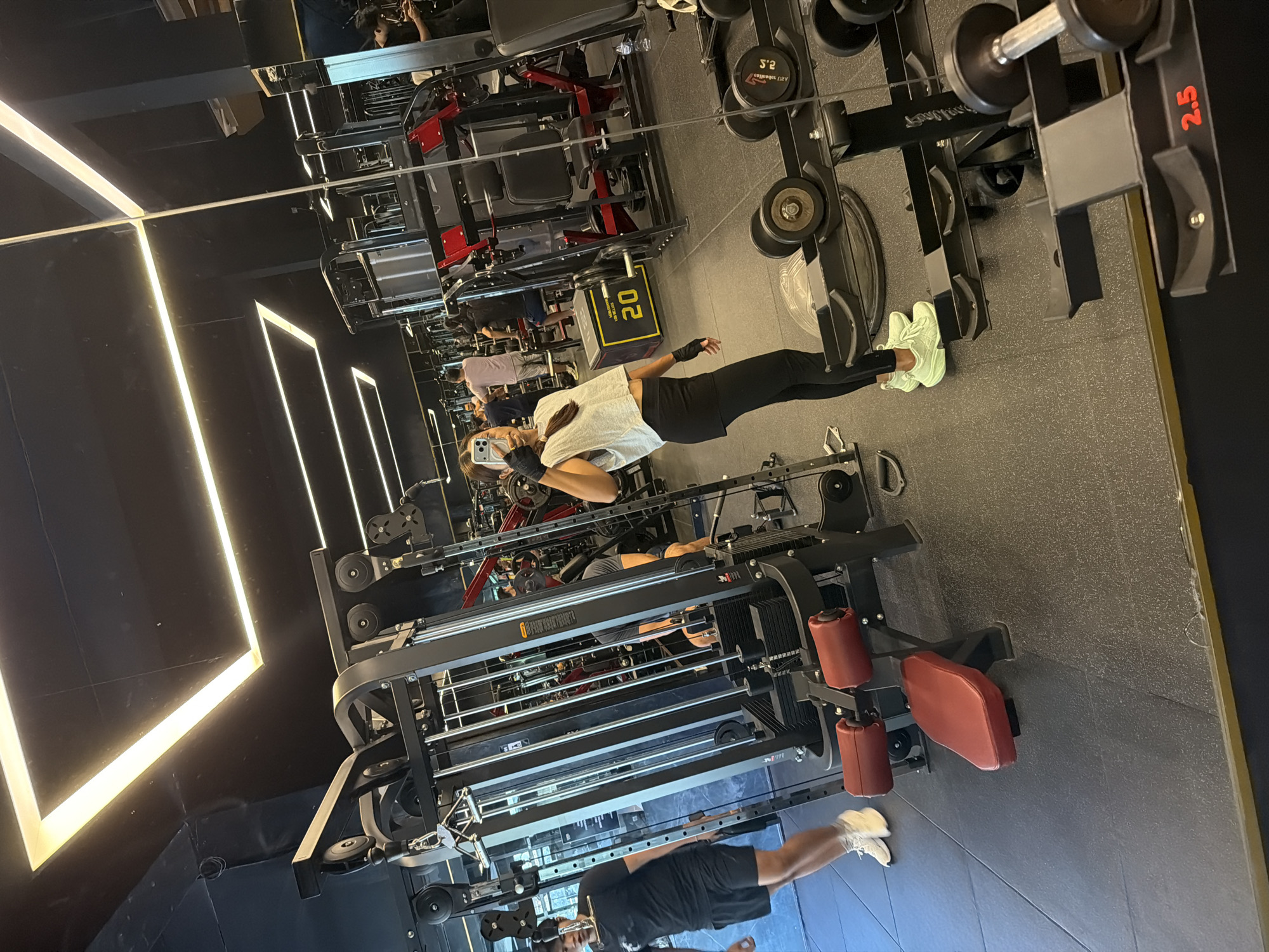 The image shows a gym interior, likely a commercial fitness center in Denpasar, Indonesia. The room is equipped with various weight training machines, including cable machines with weight stacks, dumbbell racks displaying weights, and other exercise equipment. The flooring is a dark, possibly rubberized, material. The walls are black with modern, linear LED lighting. There are two people visible, both appearing to be using smartphones, presumably taking photos or videos. One is a woman in exercise attire and the other is a man. The people are reflected in the floor-to-ceiling mirrored walls.
