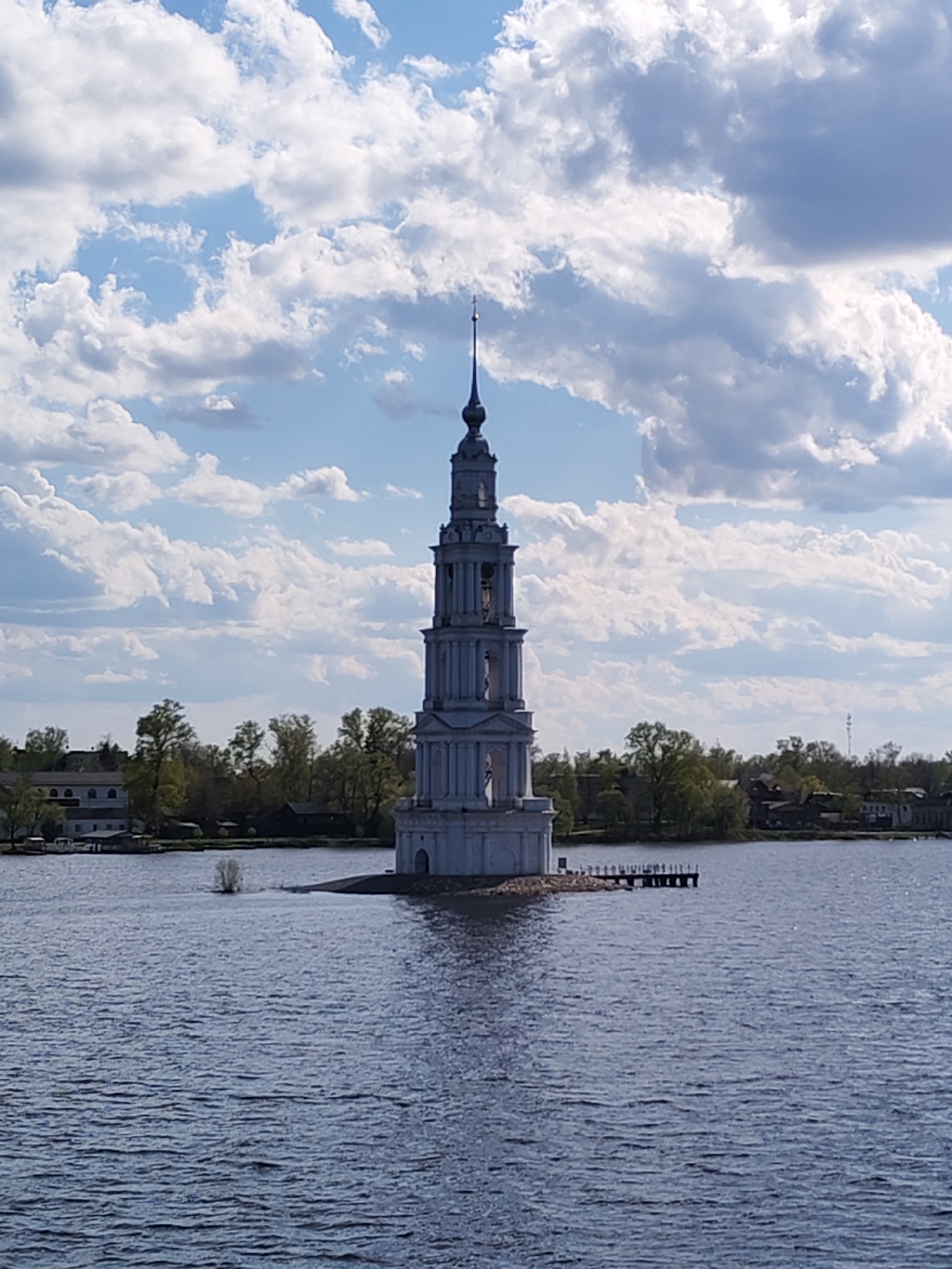 The image depicts the flooded St. Nicholas Cathedral bell tower situated in the Volga River, near the city of Myshkin, Russia. The bell tower is a tall, white structure with a spire extending into a blue sky dotted with white clouds. It stands on what appears to be a small, partially submerged island. The surrounding water is calm with gentle ripples reflecting the light. In the background, there is a visible shoreline with various buildings and trees. A small dock or pier extends from the tower into the river.