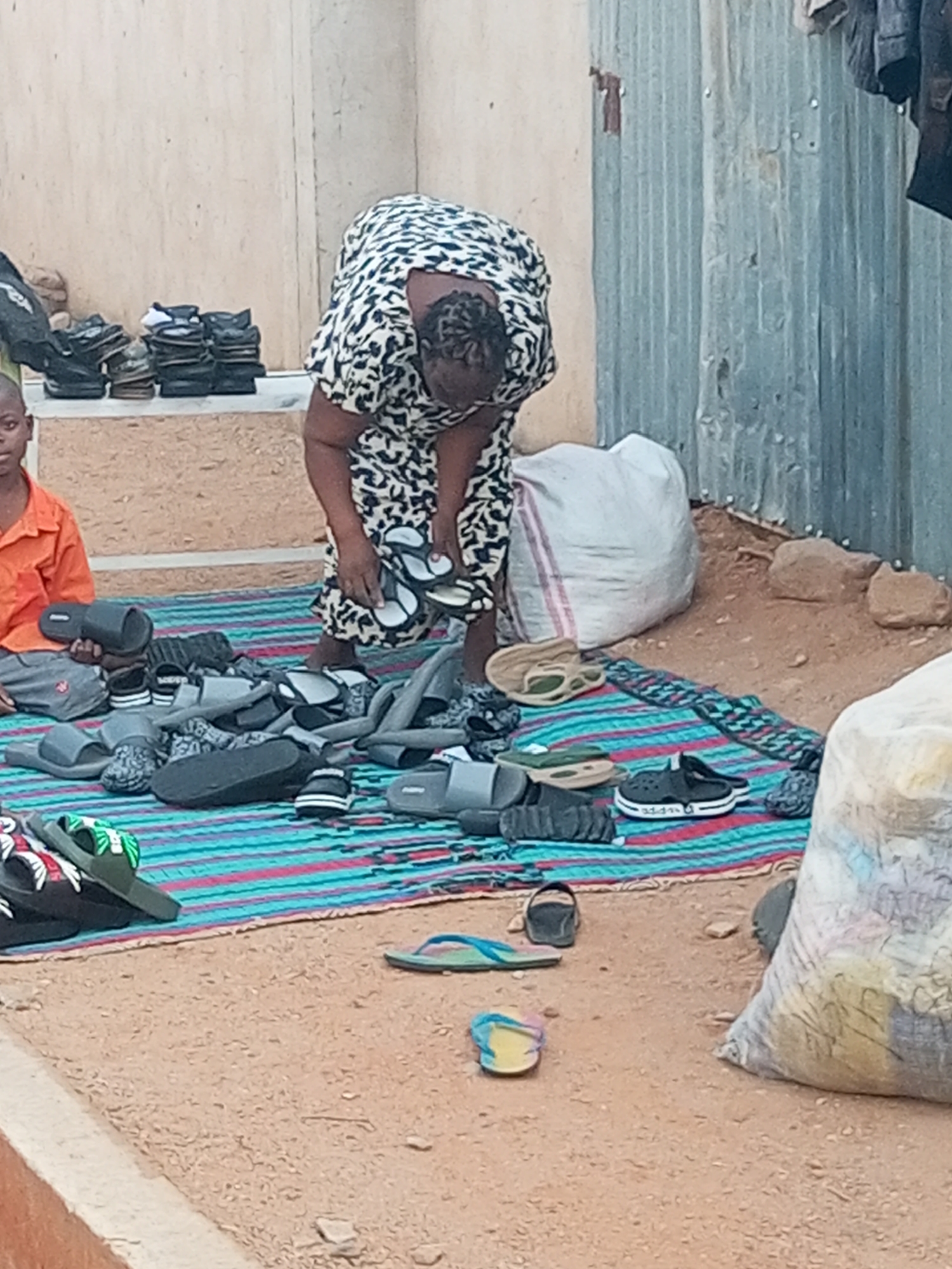 A woman in a black-and-white patterned dress stands on a blue-and-pink striped mat outdoors, surrounded by numerous pairs of sandals and flip-flops. She is bending over, holding a pair of sandals in her hands. A young boy in an orange shirt sits to her left on the mat, holding what appears to be a black sandal or slipper. Various sandals and shoes are scattered across the mat. In the background, a stack of footwear sits on a ledge. A large white bag sits behind the woman near a corrugated metal fence. Another large bag, partially visible, is in the foreground. The ground appears to be dirt. The location is identified as Tempure, Nigeria.