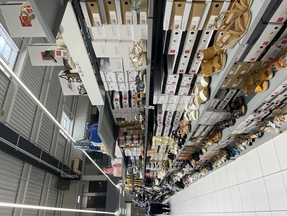 The image shows a shoe store located in Saint-Brice-Courcelles, France at the coordinates (latitude: 49.283, longitude: 4). The store's interior is brightly lit, and the walls are white. There are many shelves filled with shoes. The shelves are made of metal and are arranged in a staggered pattern. The shelves are filled with a variety of shoes, including sandals, sneakers, and boots. Some shoes are in boxes while others are displayed on shelves. There are no people in the store. The image is taken from the right side of the store, looking toward the back. The store is clean and organized. The image shows the variety of shoes for sale, including a woman's shoe on the left side of the image that appears to be on sale for 14 euros. The image is well lit and shows the store's layout. The image shows a variety of shoe styles and colors.