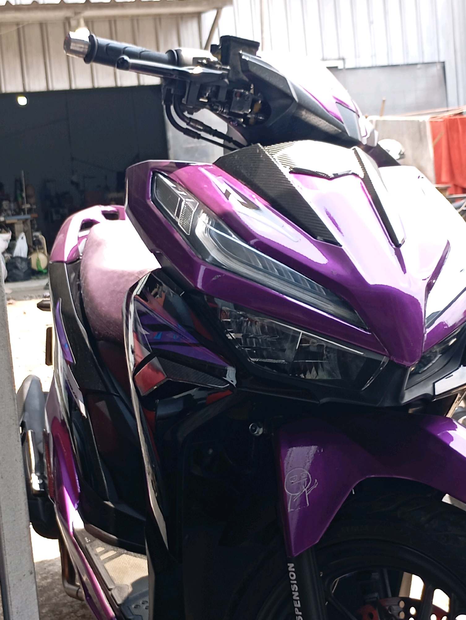 An eye-level medium shot captures the front and right side of a custom-painted purple scooter, with a portion of its dark-colored seat visible. The scooter is parked outdoors, with a building facade featuring a large roll-up door forming the background. To the left, a section of a white pillar is visible, and further back, items like motorcycles, bags, and other miscellaneous objects are partially obscured. To the right of the scooter, a red tarp is draped over an unseen object. The scooter's front fender features a small, white cartoon drawing of a figure giving the middle finger. The scooter is equipped with a carbon fiber panel on its front fairing and has "SUSPENSION" printed vertically on its front tire. The lighting suggests it is daytime. The scene is likely in an open-air workshop or parking area.