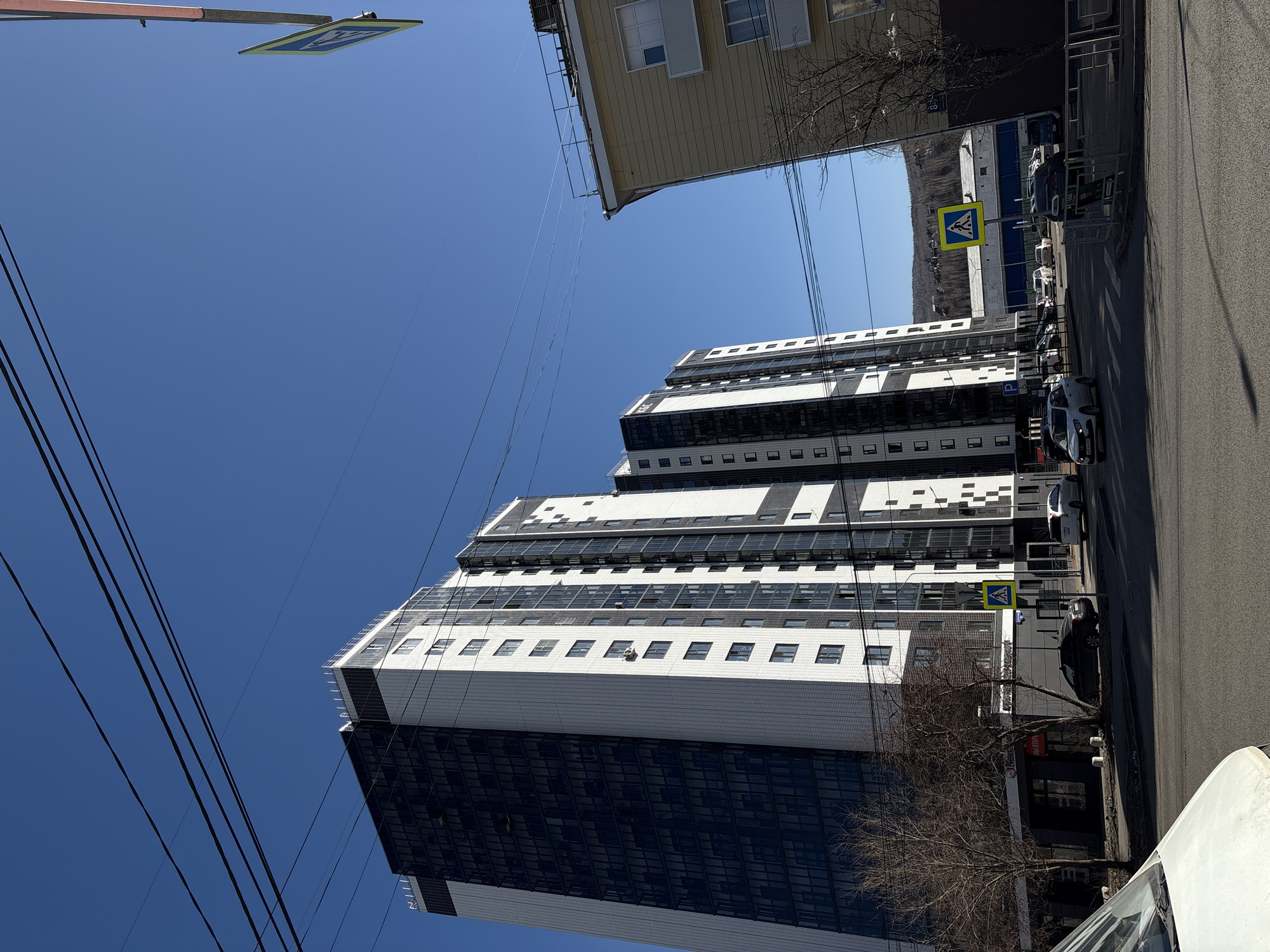 The image depicts a high-rise building in Krasnoyarsk, Russia, characterized by its modern architecture of stacked blocks with a black and white color scheme. A clear blue sky forms the backdrop. Power lines crisscross the sky. An adjacent yellow building is visible. Cars are parked along the side of the building. A pedestrian crossing sign is nearby. A bare tree sits next to the building on the street. Part of a white vehicle is in the foreground.