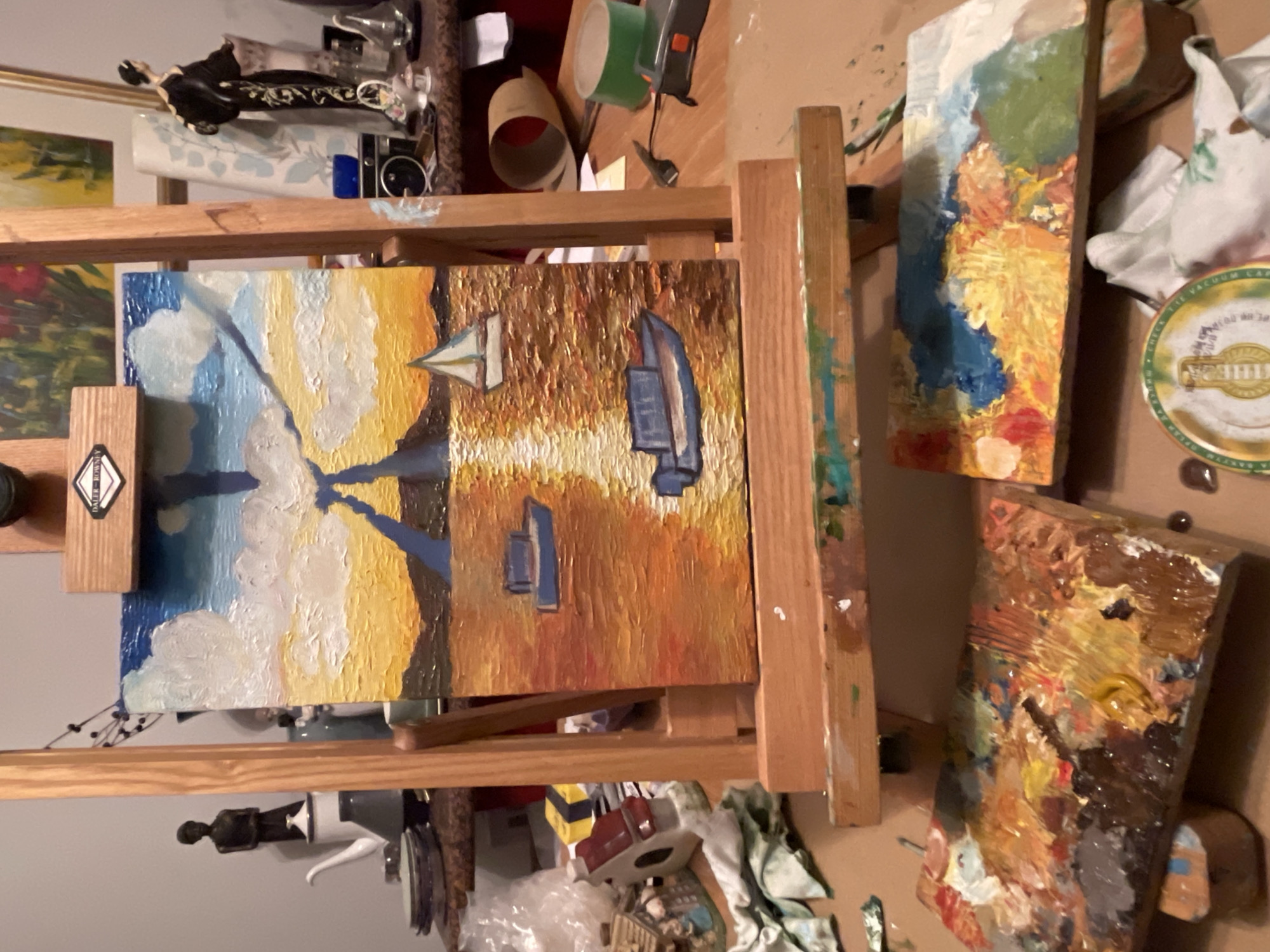 The image shows a painter's studio. An easel holds a painting of sailboats in a harbor scene. Other paintings are visible, as well as art supplies, including paint tubes, brushes, and palettes covered in paint. There is no evidence of the location being Maidstone, United Kingdom.