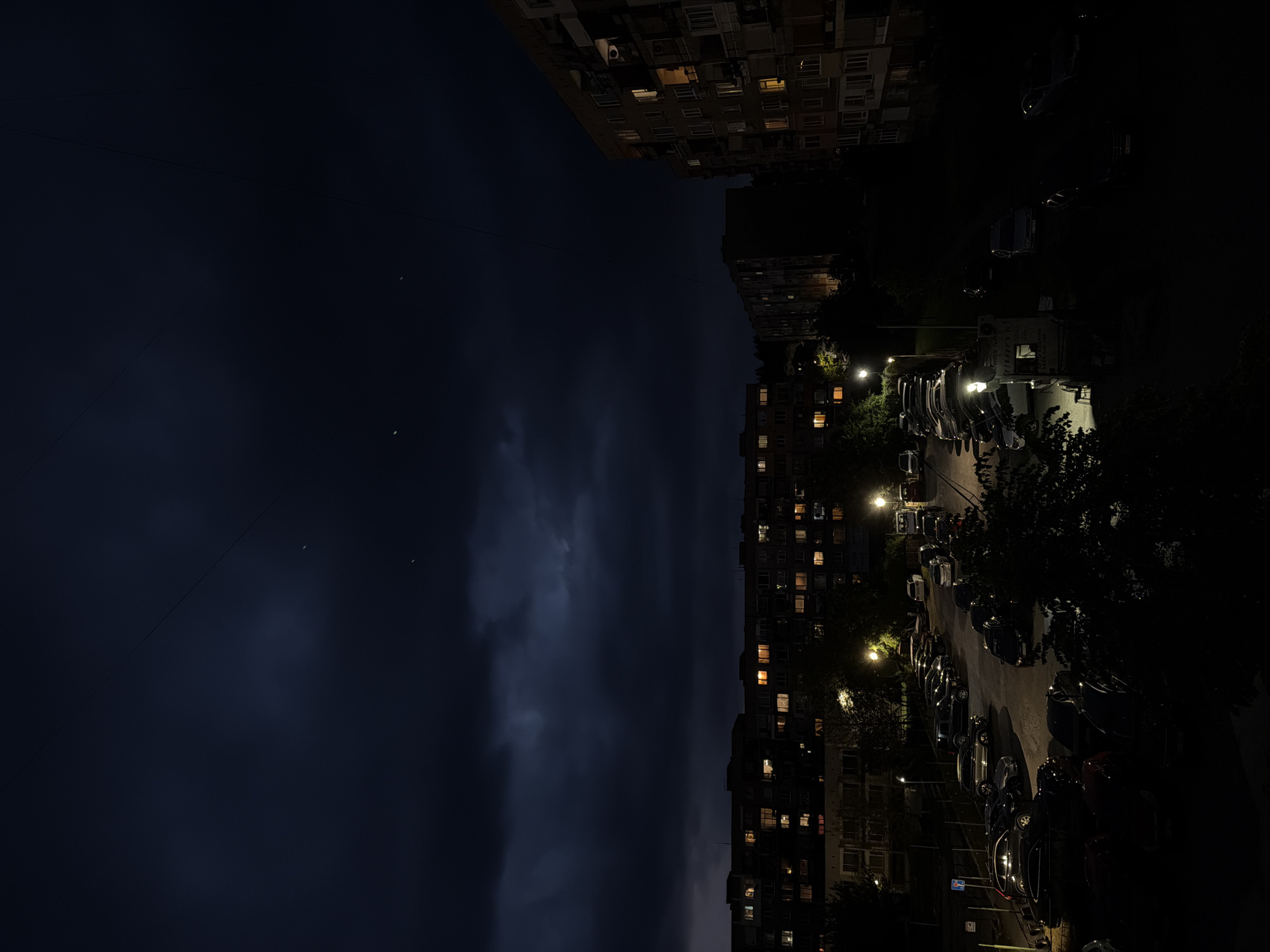 The image depicts a nighttime urban landscape. In the foreground, multiple parked cars line a street illuminated by streetlights, flanked by trees. Behind the cars, residential buildings rise, some with lit windows. The sky above is dark with cloud cover, hinting at an approaching storm. Two bright spots, likely stars or planets, are visible in the upper-left portion of the sky. The location is unknown.