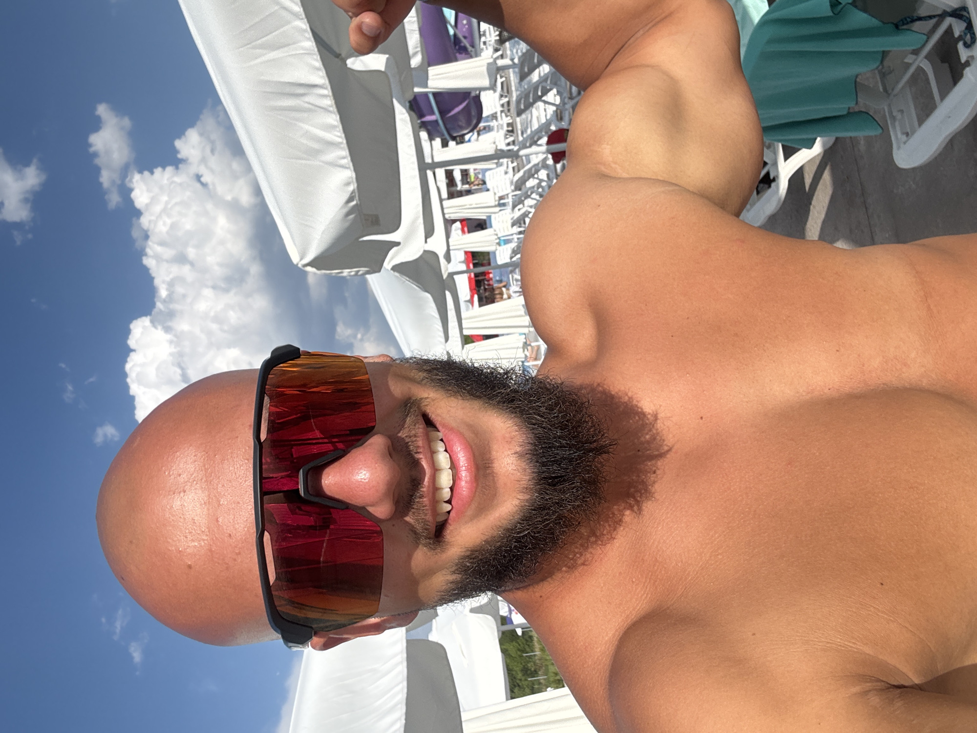 A man with a shaved head and a beard smiles at the camera, wearing red-tinted sunglasses. He is outdoors, with a blue sky and white clouds visible in the background, suggesting a sunny day. Behind him, there are white beach umbrellas and lounge chairs, and a glimpse of a purple water slide. The man is flexing his bicep, and his torso is exposed to the sun. The scene appears to be at a water park or a resort. The location is identified as Ivanyane, Bulgaria.