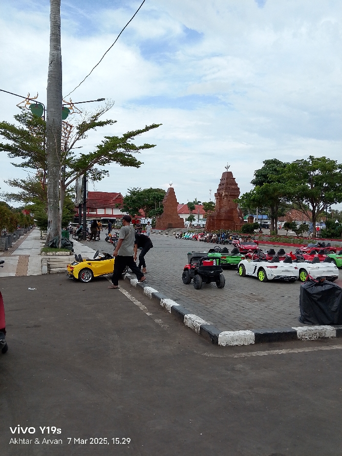 The image shows a street scene in Indramayu, Indonesia, located at coordinates -6.326 latitude and 108.322 longitude. Two men are standing on the sidewalk, one wearing a blue shirt and the other a red shirt. There are several toy cars parked in the street, including a yellow one and a white one. The street has a black and white curb. In the background, there is a brick structure, possibly a temple or monument. The sky is partially cloudy, and there are trees on either side of the street. The image appears to have been taken on March 7, 2023, at 3:29 pm.