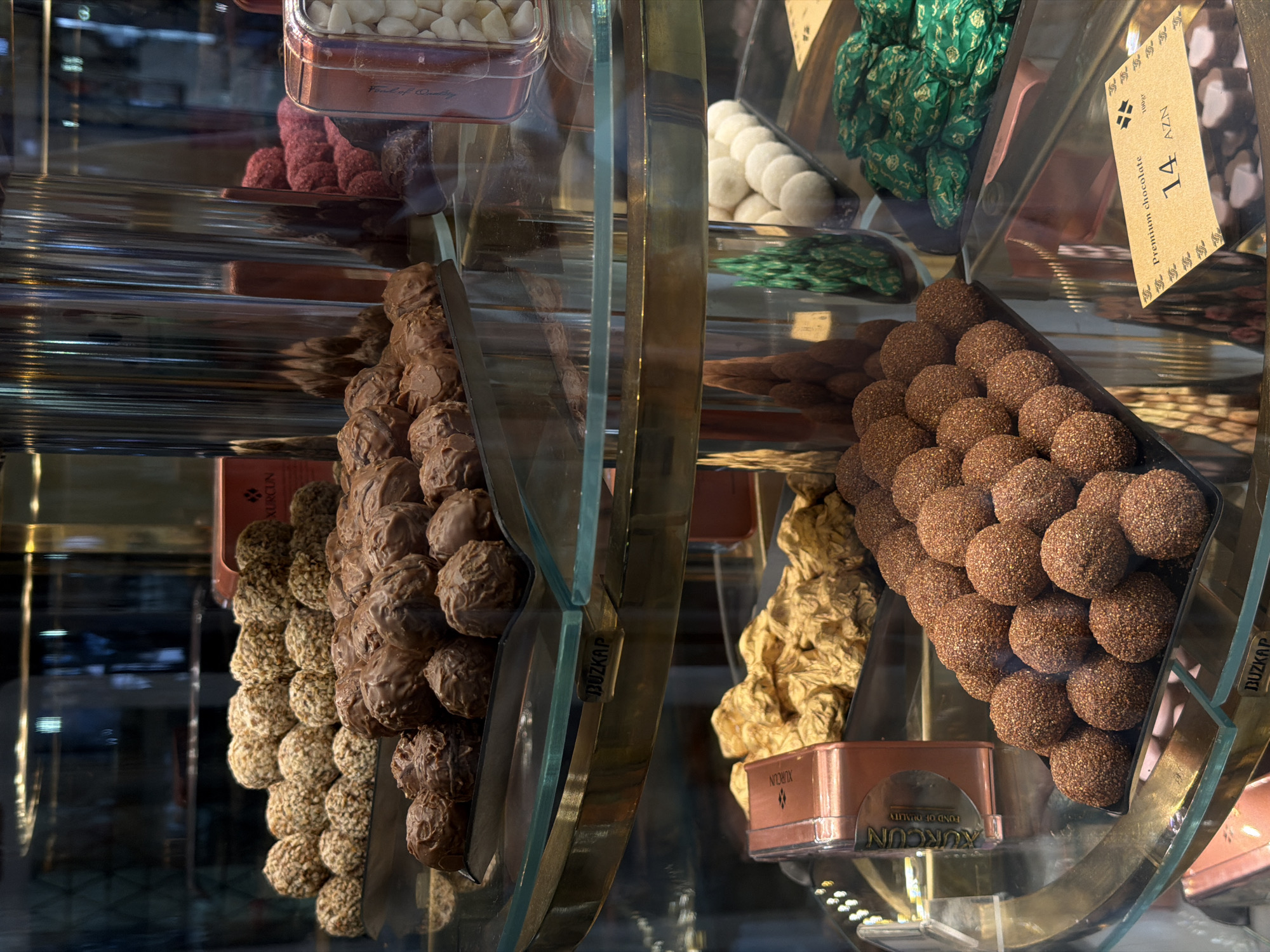 This image shows a close-up of a display case filled with chocolates and candies. The display case appears to be in a shop, possibly a confectionery or a dessert shop. In the foreground, on the bottom shelf, a tray of small, round, chocolate-covered truffles is visible. To the right of the truffles, there are several green foil-wrapped candies. A price tag indicating "Premium chocolate 14 AZN" is also visible.
On the shelf above, there are trays of various chocolates. One tray contains many light brown, irregularly shaped chocolates, and another tray displays round, light-colored candies. Behind these, there are more chocolates, including some darker brown ones. A metallic cylinder structure is also present, reflecting the surrounding items.
Further up, on the top shelf, a collection of round chocolates covered in nuts or granola is displayed. To their right, a red box with the brand name "KURCUN" is partially visible. There are also some red, spherical candies and a clear container filled with white, small, oval-shaped items. The overall impression is one of a well-stocked and attractive display of confectionery. The presence of the currency "AZN" suggests a location in Azerbaijan. Given the reference to Khazar Raion, Azerbaijan, it is plausible that this establishment is located within this administrative district of Baku, the capital city of Azerbaijan.