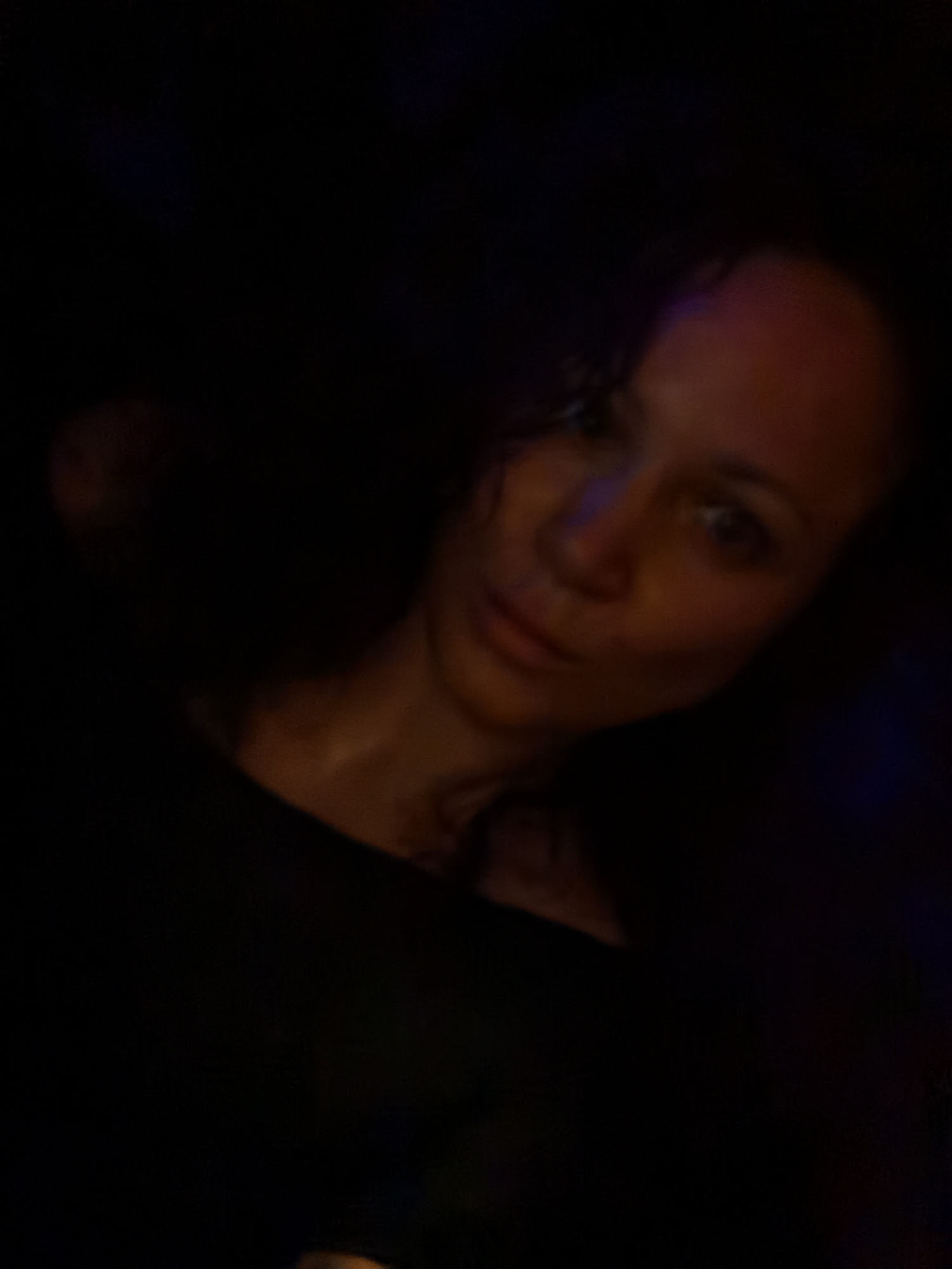 A woman with curly hair and fair skin is featured in a close-up portrait taken in a dimly lit environment. Her expression is neutral. She is wearing a dark-colored, possibly black, top. The overall scene has limited visibility due to the darkness. The location and time are unknown.