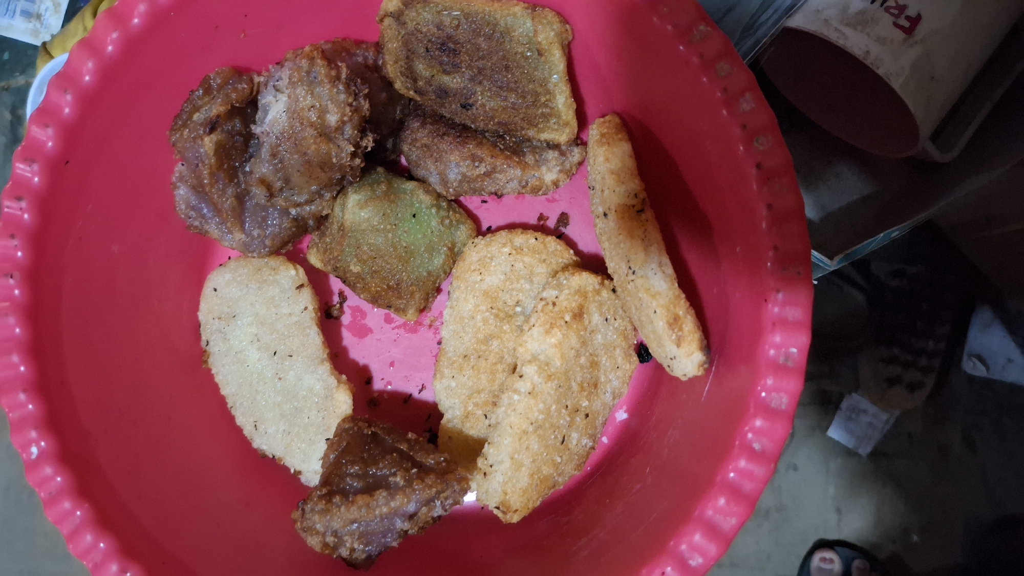 The image shows a close-up, overhead view of a bright pink plastic bowl filled with various fried food items. The food consists of several pieces of what appear to be fried dough or plantains, some light golden brown and others darker and more irregularly shaped. There are also some lighter-colored, flatter pieces that could be fried crackers or slices of food. The bowl is sitting on a dark surface, and the surrounding environment is dimly lit. In the lower right corner, part of a mug with red writing is visible. The scene appears to be domestic. The presence of the food in a bowl suggests it is being served or has recently been prepared. The writing on the mug is not fully legible but appears to start with "FR". No specific details indicate a location in Pucakwangi, Indonesia.