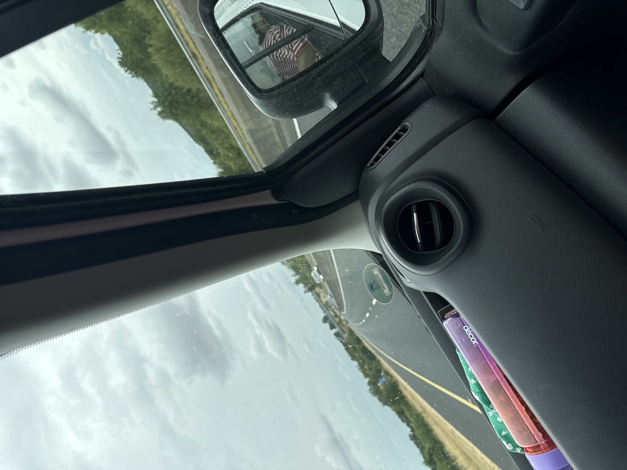 An interior view from a vehicle traveling on a highway. The dashboard is visible in the foreground, with a car vent and a few objects placed in a compartment. Outside the window, a highway stretches ahead, bordered by green trees and hills under a cloudy sky. A side mirror reflects a person wearing a striped shirt. The car appears to be in motion. The location is identified as Pill Belan, Ireland.