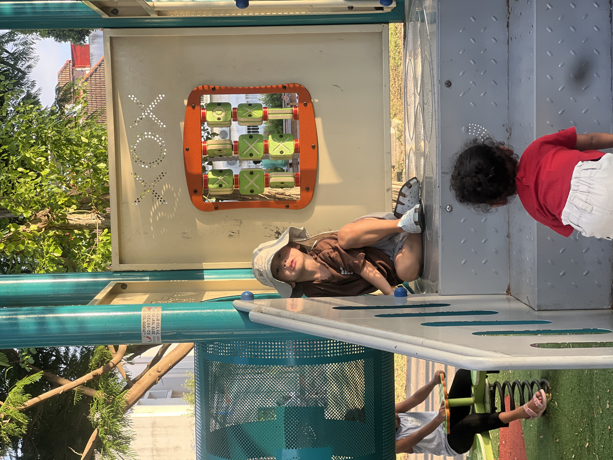Two young children are playing at a playground. The scene is outdoors on a sunny day, likely late afternoon given the warm lighting and shadows. The playground equipment is made of metal and plastic, featuring a tic-tac-toe game with green sliders on an orange frame, and a metal staircase.
One child, wearing a light-colored sun hat and a brown t-shirt, is lying on his side on a metal platform, looking up at the camera. He appears relaxed. The other child, wearing a red shirt and white shorts, is standing and looking away from the camera, near the staircase. Their ethnicity is not definitively clear.
In the background, lush green trees and buildings with red tile roofs are visible. A sticker with Hebrew text is attached to a green metal pole. The text reads: "קיאק שפשוף 02-9972005 - ת. יצור 2019 - יצרן: כל כל פלס". This translates to "Kayak scrub 02-9972005 - Production date 2019 - Manufacturer: Kol Kol Plas". This suggests the playground equipment is from Israel, consistent with the provided location context of Kiryat Bialik. The overall mood is one of casual play in a pleasant outdoor environment.