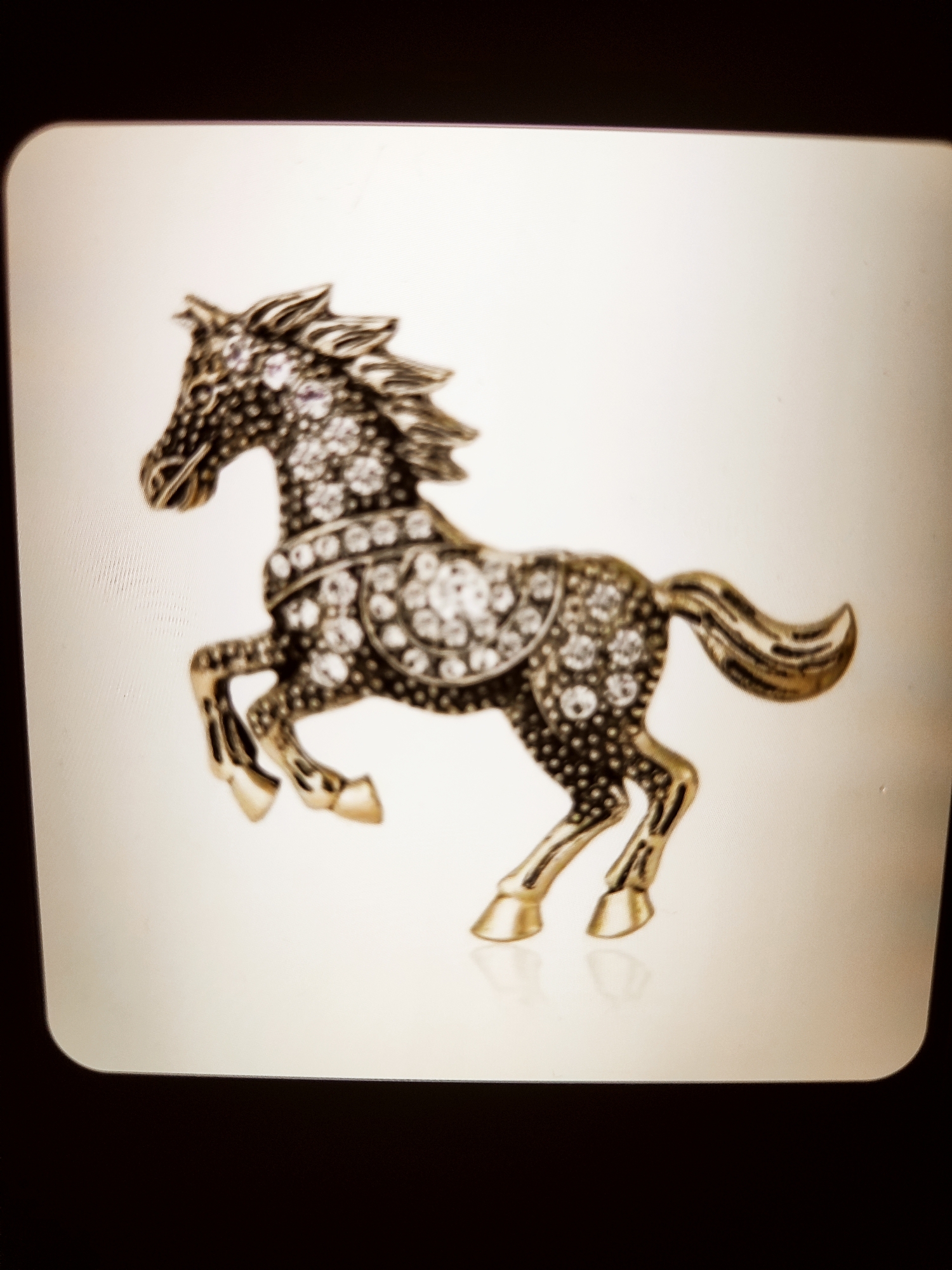 A detailed, full-body shot showcases a metallic horse brooch, adorned with numerous clear rhinestones and a few subtle purple stones. The brooch is depicted against a plain, off-white background with a slight sheen. The horse is in a dynamic pose, with its front legs raised as if in mid-gallop, and its tail flowing elegantly. The metallic finish suggests a gold or bronze tone, intricately detailed to capture the musculature and mane of the animal. The backdrop is featureless, offering no environmental context, therefore the location cannot be determined.