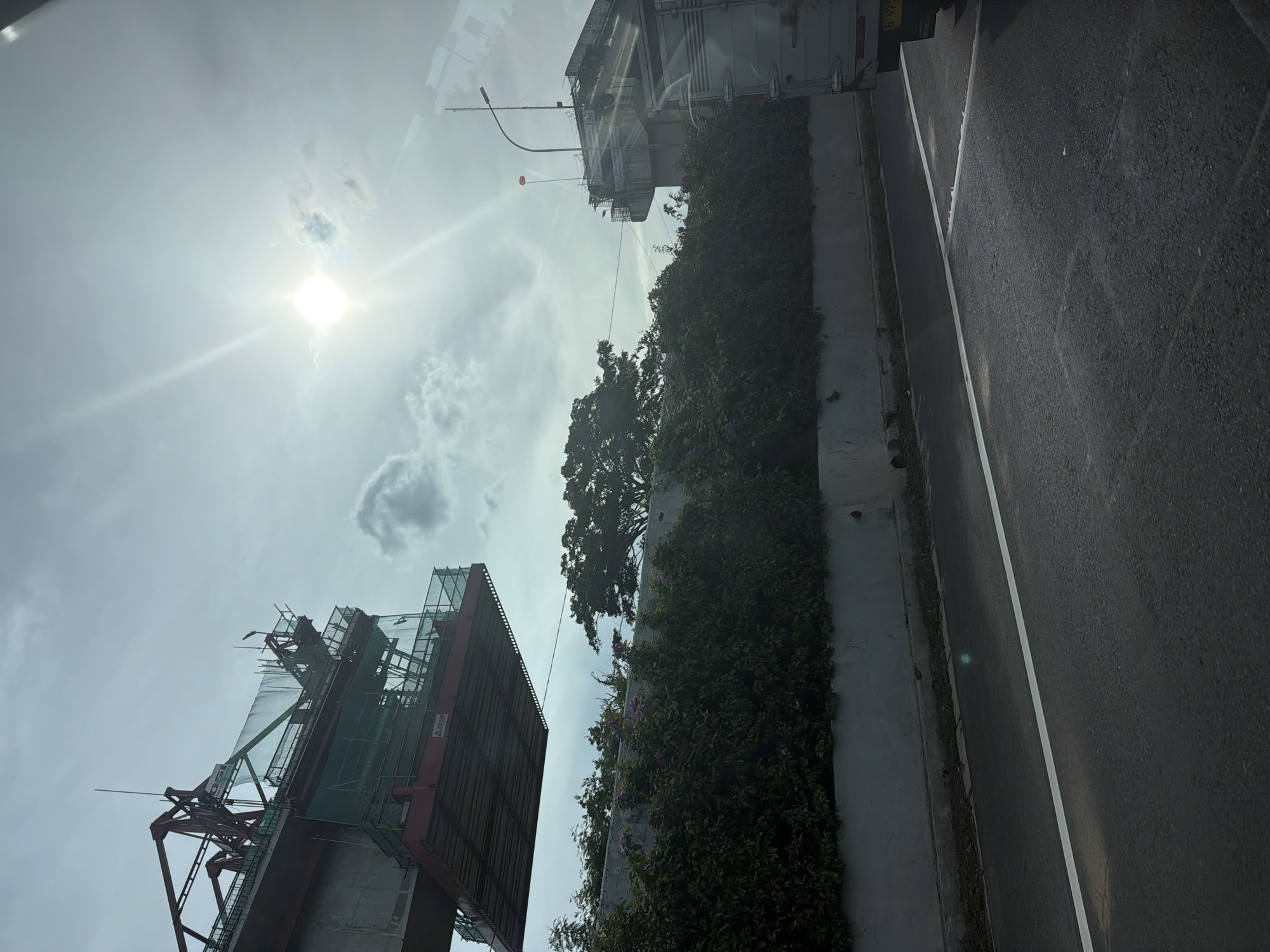 The image depicts a scene in North Jakarta, Indonesia. The view is angled upwards towards the sky, which is partially obscured by clouds and sunlight. Visible landmarks include a construction site with scaffolding and a building covered in vegetation. A road with a white lane marking is present on the right side of the frame. A truck is visible in the background near the building. No people are visible in the image.