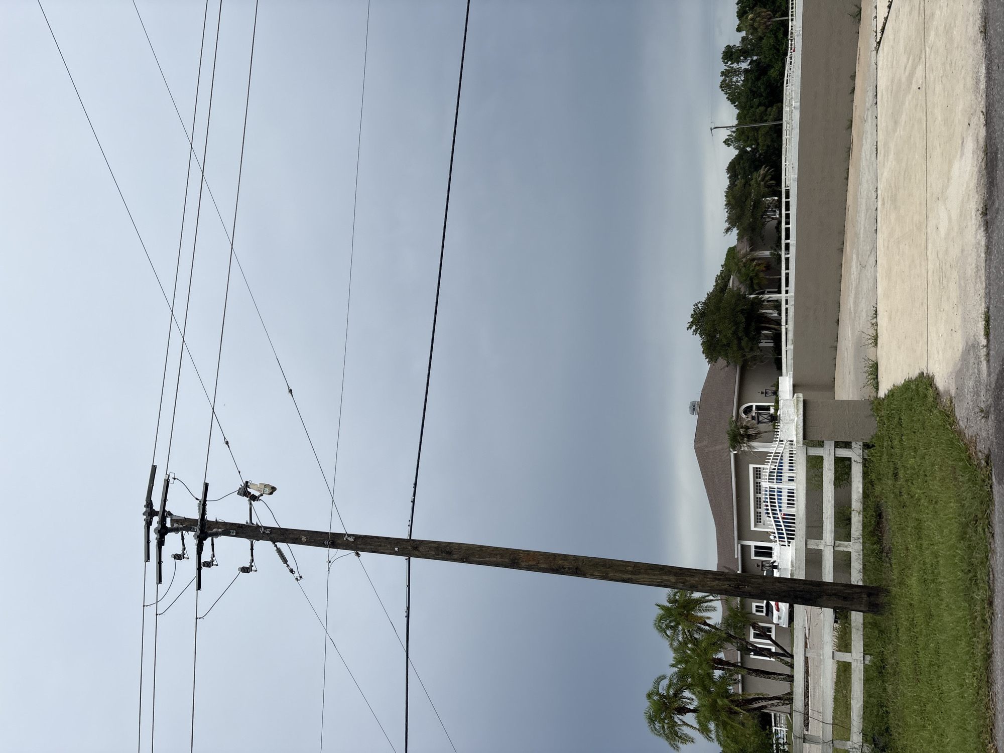 The image captures a street scene in Bradenton, Florida, United States. A prominent wooden utility pole with electrical wires stands in the foreground, leaning slightly to the left. Beyond the pole, a large, light-colored house with a grey roof is visible, featuring multiple windows and a garage with a blue gate. Palm trees and other greenery surround the property, and a white fence with decorative posts delineates the front yard. The sky is overcast with heavy grey clouds, suggesting a potentially stormy day. A paved driveway leads up to the house. No people are visible in the scene.