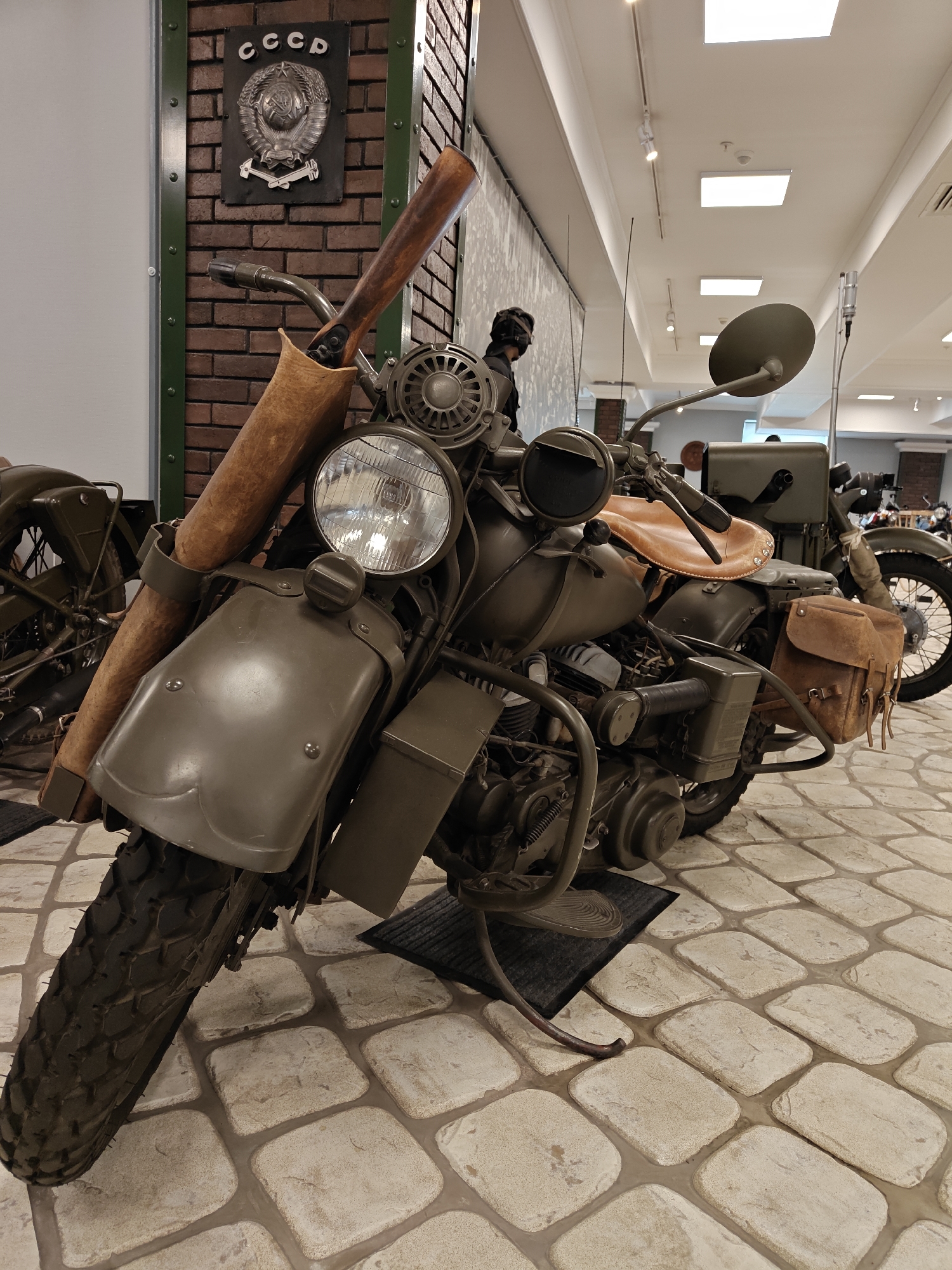 The image shows a vintage military motorcycle on display in what appears to be a museum or showroom. The motorcycle is olive green and has a sidecar. A rifle is mounted on the front. A CCCP plaque is visible on a brick wall in the background, suggesting a Soviet-era origin. The floor is paved with stone tiles. The presence of other motorcycles in the background indicates a collection or exhibit. Based on the information provided, the location is the city of Архангельское, Russia.