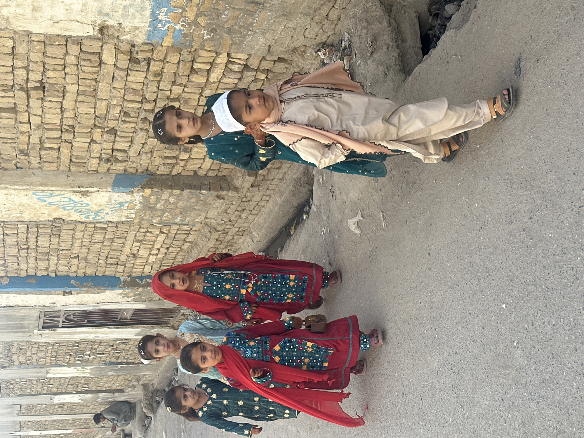 In Quetta City Tehsil, Pakistan, six children are captured outdoors on an overcast day. The setting appears to be a street adjacent to a brick building with a partially visible window and aged facade. The children are dressed in traditional attire, with three wearing green and others in red dresses. The two eldest are wearing more formal garments. A man is partially visible in the lower left corner, crouching down. The ground surface is paved, suggesting a walkway or road.