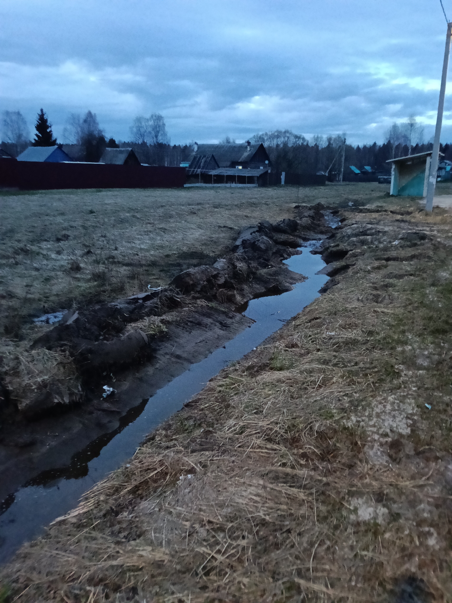The image depicts a drainage ditch filled with water, located in a rural or semi-rural area. The ditch runs through a field of dry grass and dirt, and the banks of the ditch are eroded. In the background, there are several buildings, including houses with varied rooflines, and a red fence. A small, dilapidated structure is visible on the right side of the image, near a utility pole. The sky is overcast, suggesting it is either early morning or late evening. There are no people visible in the image. The general location appears to be a residential area within a countryside setting. The exact location of Unknown, Unknown cannot be determined.