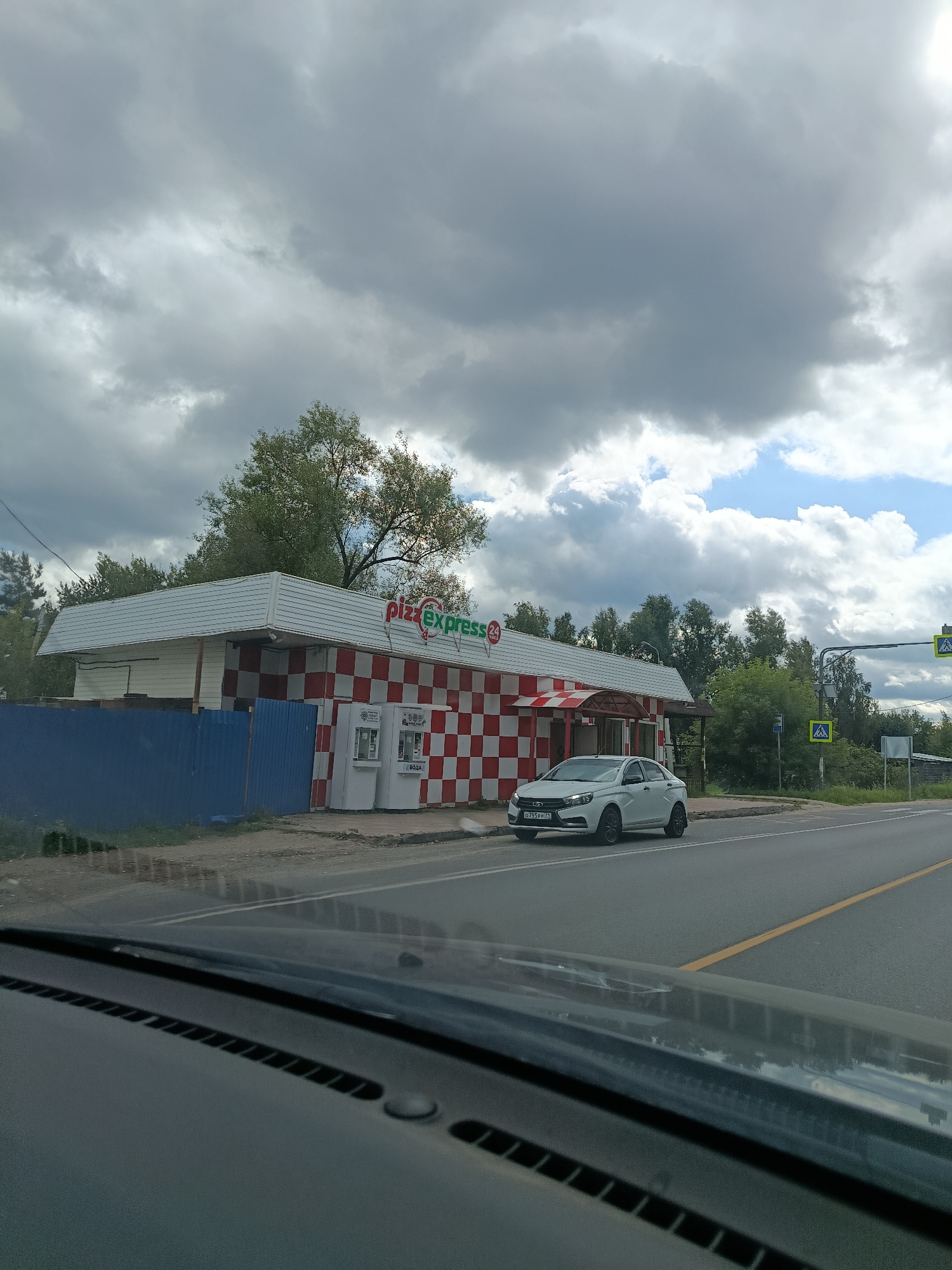 The image shows a white sedan parked in front of a Pizza Express establishment in Снопок Новый, Russia. The building features a checkered red and white facade. Two vending machines are visible next to the entrance. A blue metal fence runs along the property line. The street in front of the building is paved with asphalt and has yellow lane markings. Pedestrian crossing signs are visible on the right side of the road, along with a traffic light. The sky is cloudy, with patches of blue sky visible. Trees and foliage are present in the background. The photo appears to be taken from inside a vehicle, looking out through the windshield.