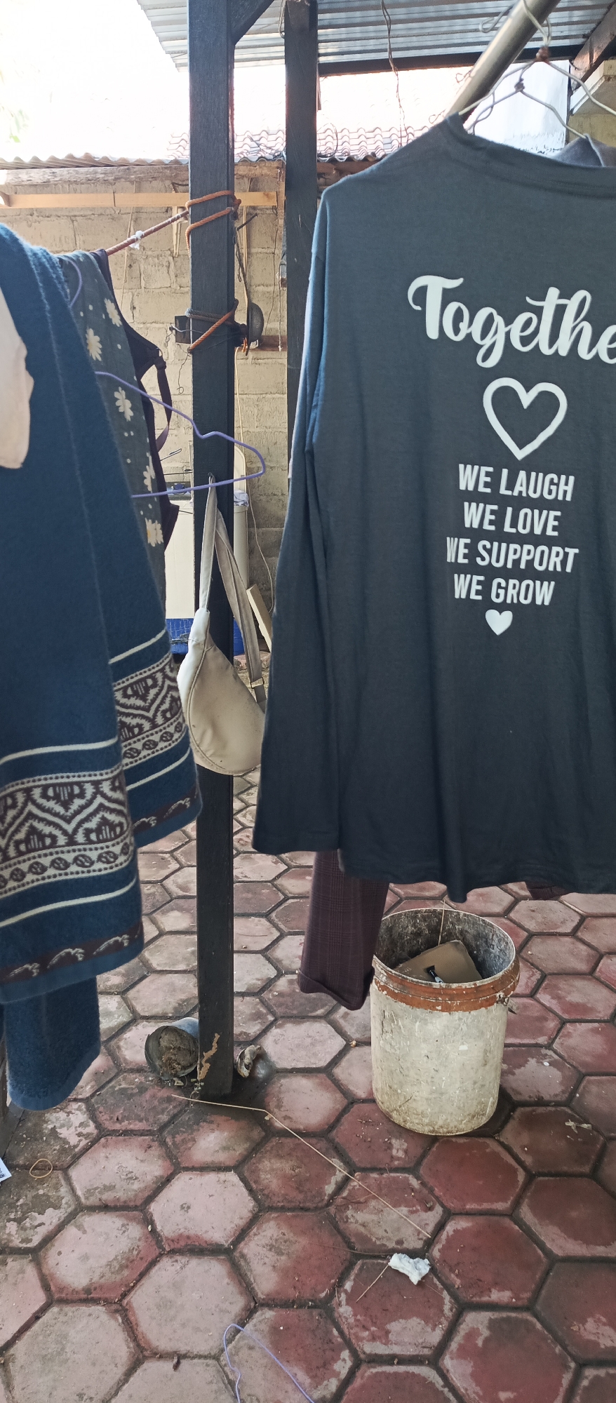 In Arjasa, Indonesia, a long-sleeved t-shirt with white text and a heart graphic is hanging on a clothesline. The text on the shirt reads "Together We Laugh We Love We Support We Grow" with two heart graphics. To the left of the t-shirt, a blue garment with a decorative pattern is also hanging. A light-colored handbag is suspended from a purple hanger. The scene is set outdoors, with a rustic, tiled floor composed of hexagonal red and grey tiles. A weathered bucket sits on the ground near the hanging clothes. The background shows a brick wall and a corrugated roof structure, suggesting a residential or outdoor service area.