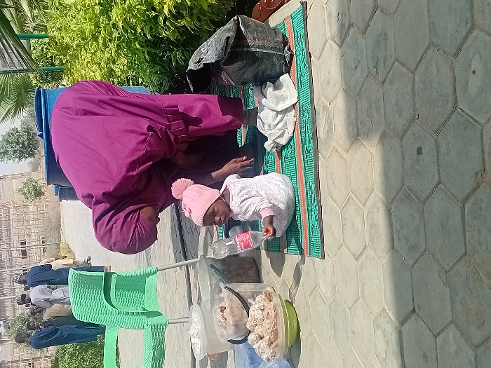 An outdoor scene in Kwazailewa, Nigeria, depicts a woman and a child on hexagonal paved ground. In the foreground, a young child, dressed in a white patterned long-sleeved garment and a pink knitted hat with a pom-pom, sits on a green and orange woven mat. The child holds a clear plastic bottle with a red label in its left hand and looks towards the camera. To the child's right, a crumpled white cloth and a dark fabric bag are present on the mat. A woman wearing a deep purple hijab and a matching long garment is bent over the mat, facing left, with her back partially towards the camera, appearing to attend to the child or items on the mat.
To the left of the mat, a partially visible light green plastic chair stands near several containers: two clear plastic buckets containing various items (one with white, fluffy material, another with brownish, irregularly shaped contents), and a light green bowl holding numerous spherical, light-colored food items. In the mid-ground, behind the woman, two large blue plastic barrels are visible. Further in the background, a group of approximately five to six men, dressed in various traditional attire, stand conversing on a paved path. Behind them, a large, multi-story building under construction, primarily concrete framing, is visible. The scene is illuminated by strong sunlight, casting distinct shadows. Green bushes and plants are present on the right and left sides of the image, with palm fronds visible at the top right.