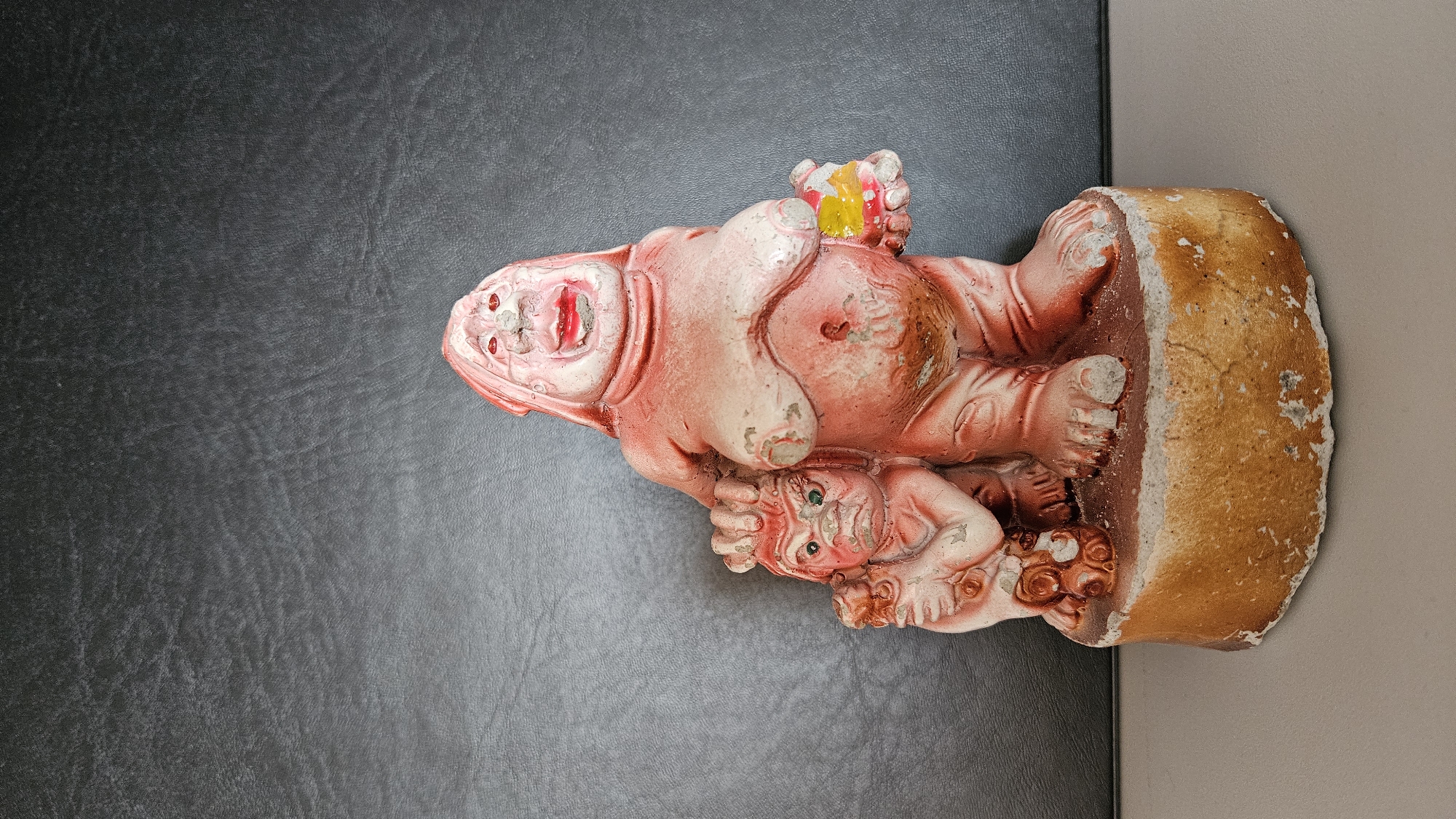 The image displays a weathered, reddish-pink statue depicting two figures laid horizontally. The larger figure, occupying the upper left portion of the statue, is robust and appears to be an adult. Its head faces upwards with an open mouth, revealing red lips and a suggestion of teeth, conveying an impression of laughter or a loud expression. The figure's body shows considerable wear, with chipped paint exposing a lighter, underlying material and areas of yellowish-brown discoloration.
A smaller, child-like figure is nestled beneath the larger one, positioned towards the bottom of the statue. This figure is also reddish-pink with distinct green eyes, looking upward, resting close to the larger entity in a pose suggesting endearment or protection.
The statue rests on a roughly textured, brownish-beige base, which appears to be made of concrete or a similar heavy material. This base is situated on a dark grey surface, possibly textured fabric or leather, which occupies the left and central portion of the background. To the right of the statue and its base, a plain light beige background is visible, likely a wall. The lighting is diffused and even, highlighting the aged details and worn textures of the statue. No text is visible in the image. The overall appearance of the statue suggests it is an antique or a well-worn decorative piece.