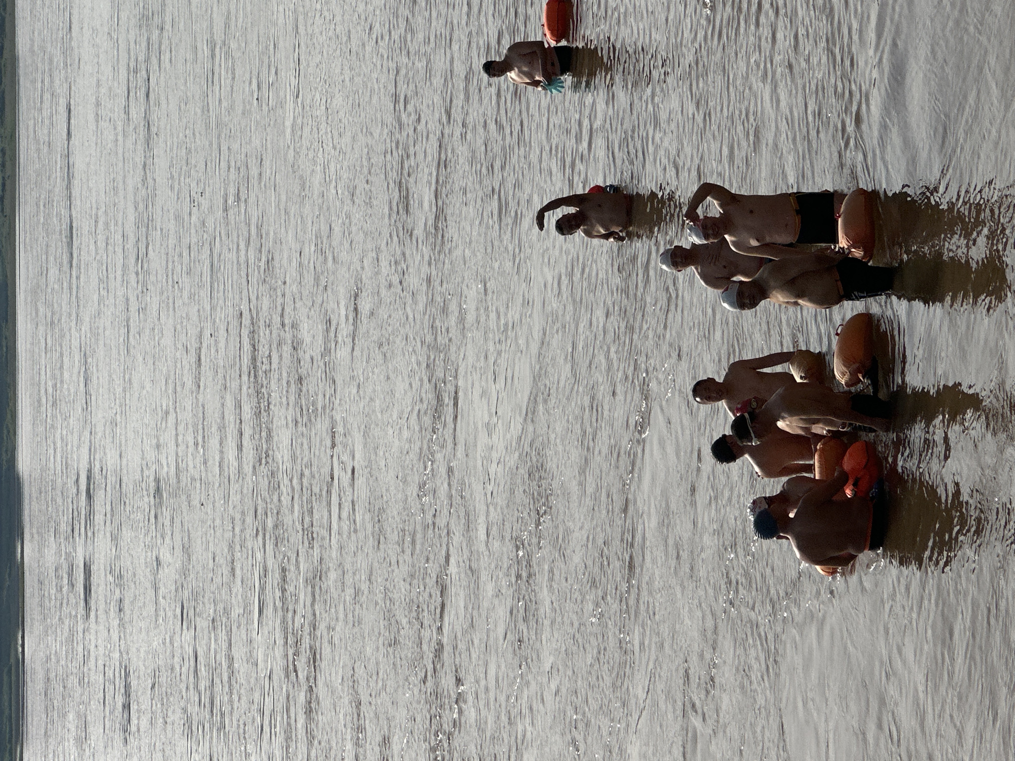 A group of approximately seven men is captured in an open body of water, likely a river, consistent with the location context of Jiangjin, China. The image is vertically oriented, with the left two-thirds dominated by the textured surface of the water, showing shimmering sunlight reflections and numerous ripples.
The men are positioned on the right side of the frame, standing or wading chest-deep in the water. They are shirtless, wearing swimming trunks, and several have white or dark swimming caps. Each swimmer appears to be accompanied by a bright orange inflatable swim buoy, typically used for visibility and safety in open water. One man at the top right of the group also wears light blue gloves.
The individuals seem to be preparing for or pausing during an open-water swim, looking towards the bottom right of the frame. The strong, directional sunlight suggests either early morning or late afternoon, creating highlights on the water and the swimmers' skin, and casting soft shadows. The overall atmosphere indicates an organized or communal swimming activity. No visible text is present in the image.