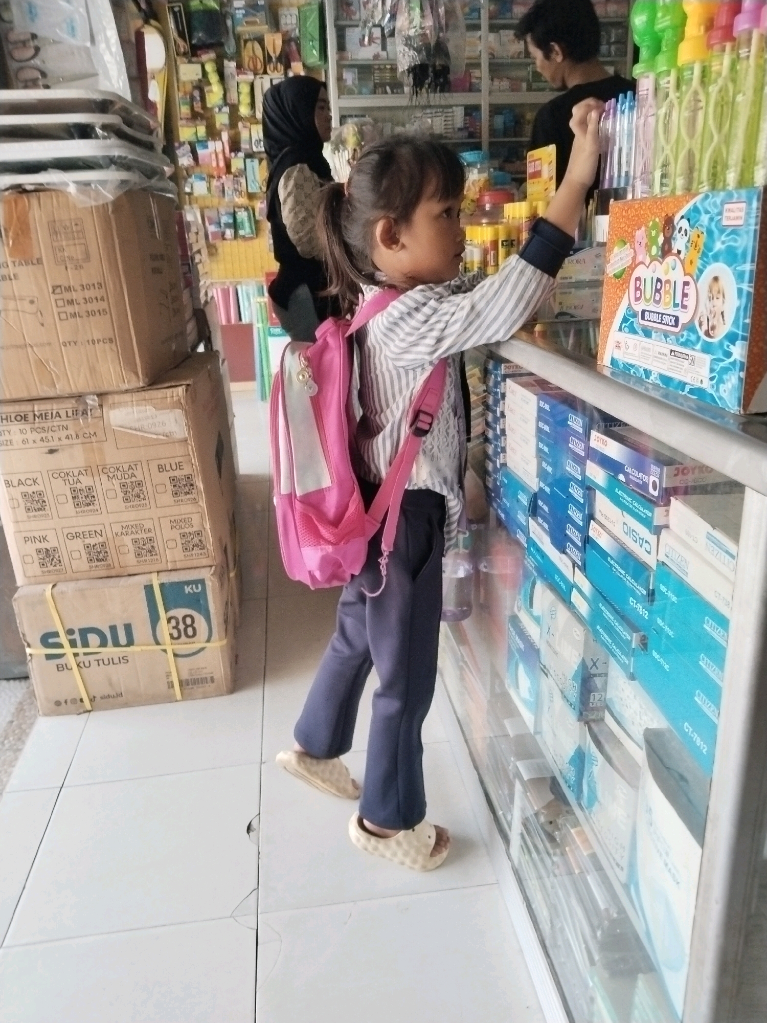 A young girl wearing a striped long-sleeved shirt, navy pants, and beige slip-on sandals stands in a store, reaching for items on a display shelf. She has a pink backpack on. In the background, a woman wearing a black hijab and a patterned shirt is visible, and a man in a black shirt stands behind a counter. The store appears to be a stationery or general goods shop, with shelves stocked with various products including stationery boxes, calculators, and bubble sticks. The floor is tiled. The context implies the location is within the city of Serang, Indonesia, based on the presence of the "Sidu" brand on a box, which is a prominent Indonesian stationery manufacturer.