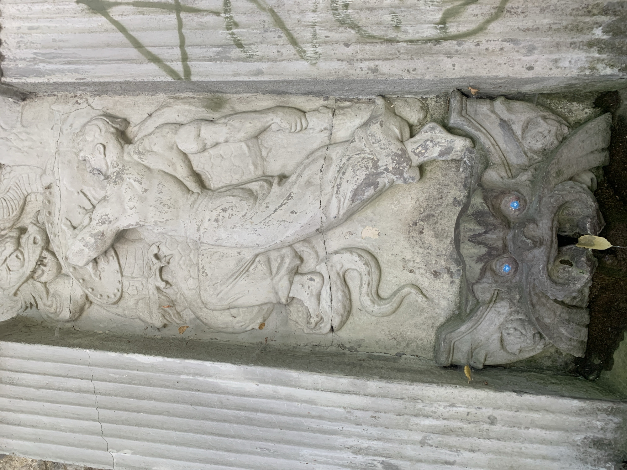 This is a medium shot of a sculpted relief on a weathered stone structure in Lviv, Ukraine. The relief depicts a mythological or historical scene with a human figure in dynamic action. The human figure, rendered in a classical style, appears to be engaged in a struggle with a serpentine creature, part of which is visible coiled around the figure's leg. Above the human figure, a more monstrous head, possibly a dragon or a demon, is partially visible. Below the main relief, a gargoyle-like spout with menacing facial features and blue glass eyes is integrated into the structure, suggesting it may be part of a fountain or water feature. The stone is heavily textured and shows signs of age, with cracks, chips, and staining. The surrounding environment appears to be outdoors, with a rough, textured wall on the left and a stained, concrete-like surface with faint green markings on the right.