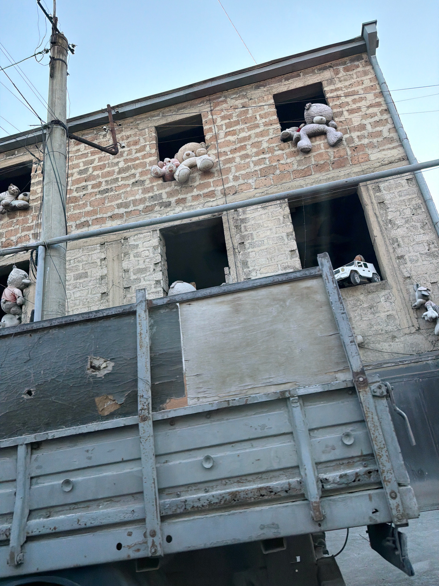 The image captures a dilapidated brick building in Yerevan, Armenia, with numerous stuffed animals placed in its open windows. The building appears to be abandoned or under construction, with exposed brickwork and missing window panes. A utility pole with wires stands prominently in the foreground, adding to the urban decay aesthetic. A large, rusted truck bed is positioned in front of the building, obscuring part of the lower facade and further emphasizing the scene's dishevelment.
The stuffed animals, primarily teddy bears of various sizes and colors, are arranged in a seemingly haphazard manner, with some peering out of the windows, others perched on ledges, and one even appearing to be tied to a wire. Their presence creates a surreal and melancholic atmosphere, hinting at a past occupancy or perhaps a form of artistic expression in an otherwise neglected urban space. The overall impression is one of abandonment, resilience, and a touch of the uncanny.