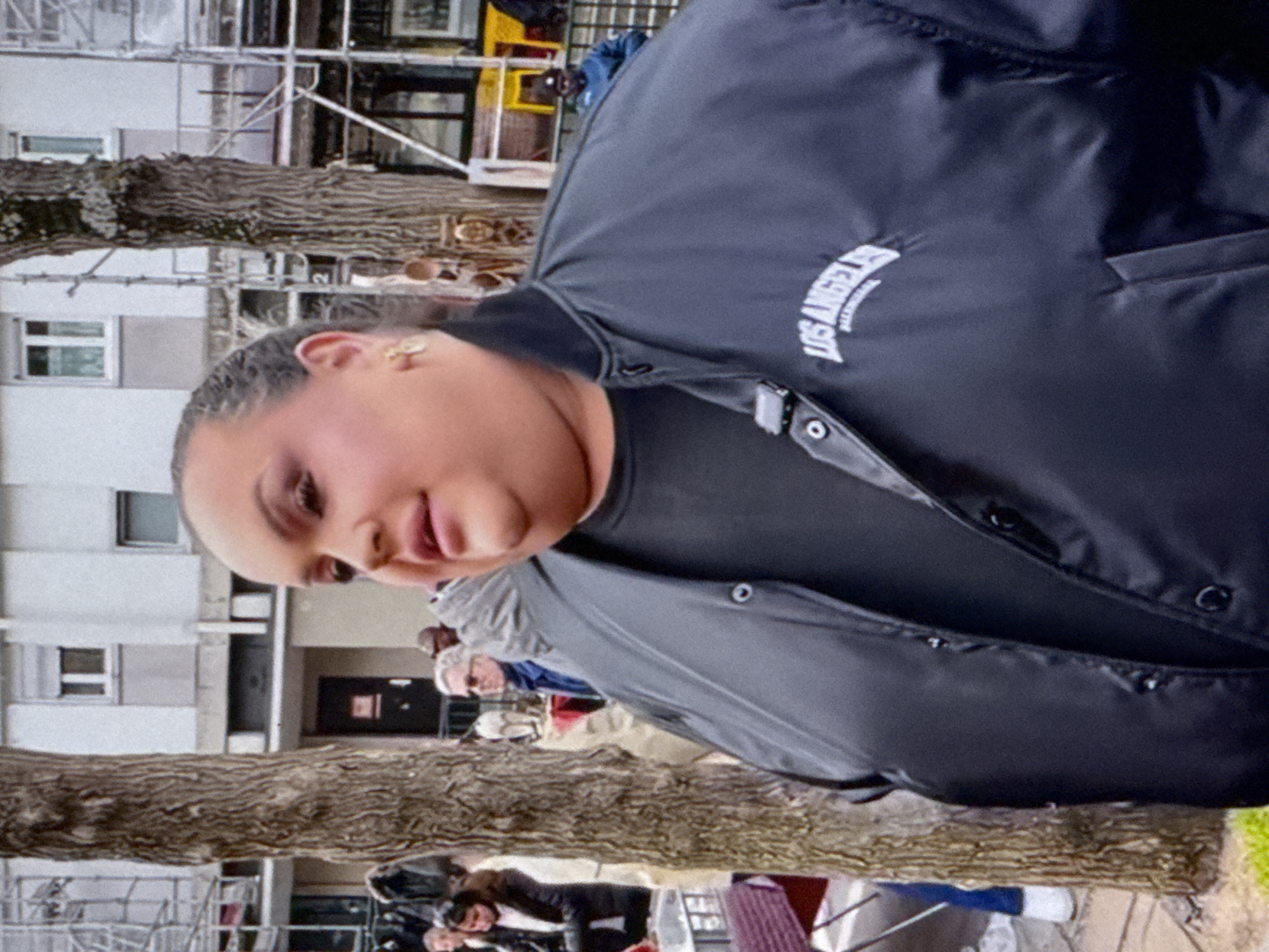 The image displays an outdoor urban scene, located in North Kuta, Indonesia. In the foreground, a woman is captured from the chest up, turned slightly to her right with a neutral to subtly pleasant expression. She is wearing a dark, possibly black, jacket that features a prominent white, arched "LOS ANGELES" text on the chest, beneath which smaller, illegible text is visible. A small, gold-colored stud earring adorns her right ear, and her dark hair appears to be pulled back.
Behind the woman, two thick-trunked trees with textured bark frame the mid-ground. In the background, several out-of-focus individuals are visible. The setting is dominated by multistory buildings, which feature numerous windows and balconies, suggesting a residential or commercial district. Scaffolding is present on one of the background structures, indicating ongoing construction or maintenance. The scene is illuminated by natural daylight, suggesting either an overcast day or soft afternoon lighting conditions.