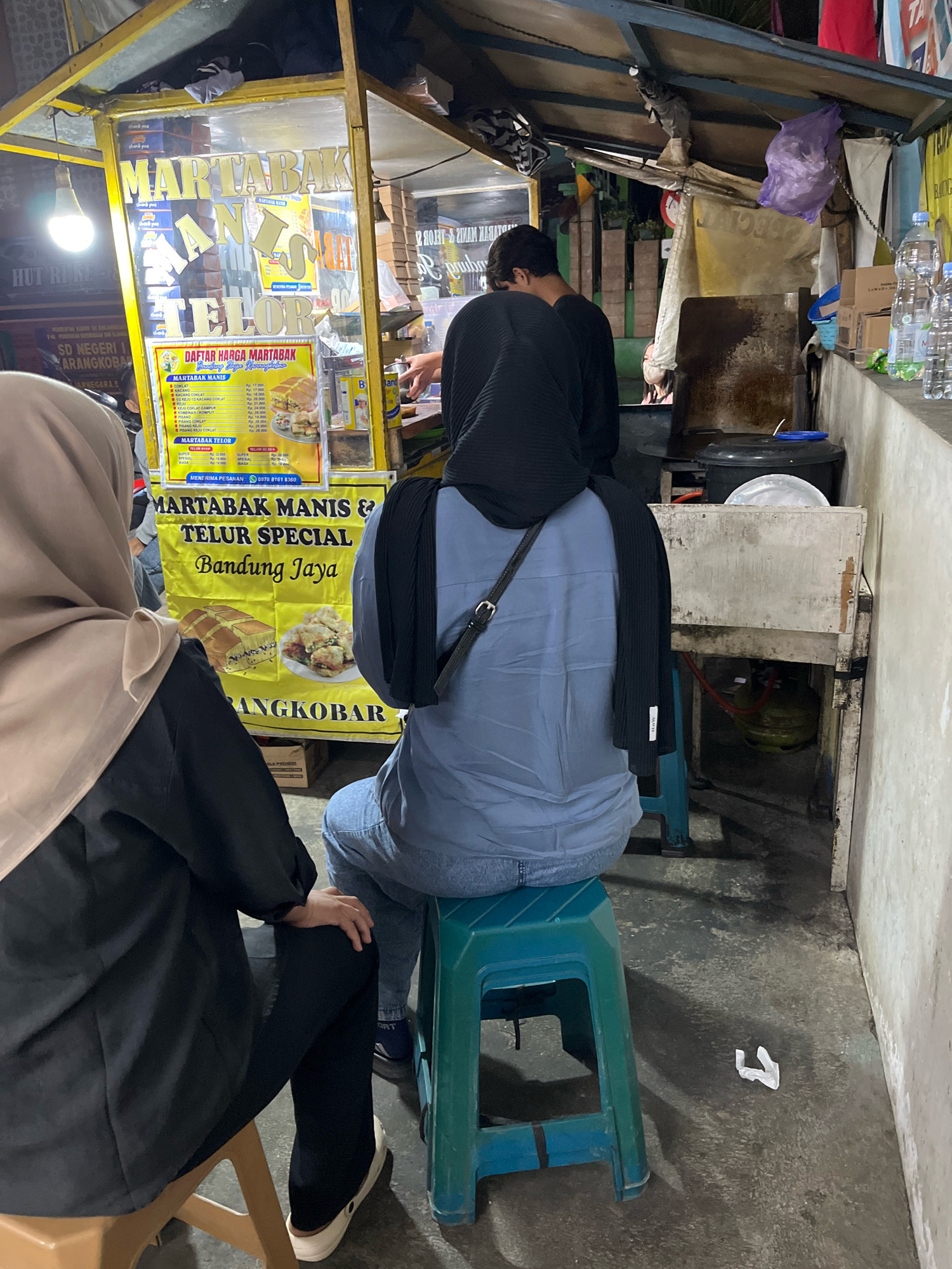 The image depicts an outdoor food stall in Karangkobar, Indonesia, specializing in martabak. Two individuals, seen from the back, are seated on stools. One person, wearing a beige hijab and a black outfit, sits on a light brown stool on the left. The other person, with a black hijab and a blue shirt, is seated on a green stool in the center of the frame. A part of the martabak stall is visible, with a yellow sign displaying "MARTABAK MANIS & TELUR SPECIAL Bandung Jaya" and a price list for martabak. Behind the stall, a gas cylinder is visible. The environment appears to be a street or alleyway, with concrete flooring and a wall on the right side. Several bottles of water are also placed on a ledge.