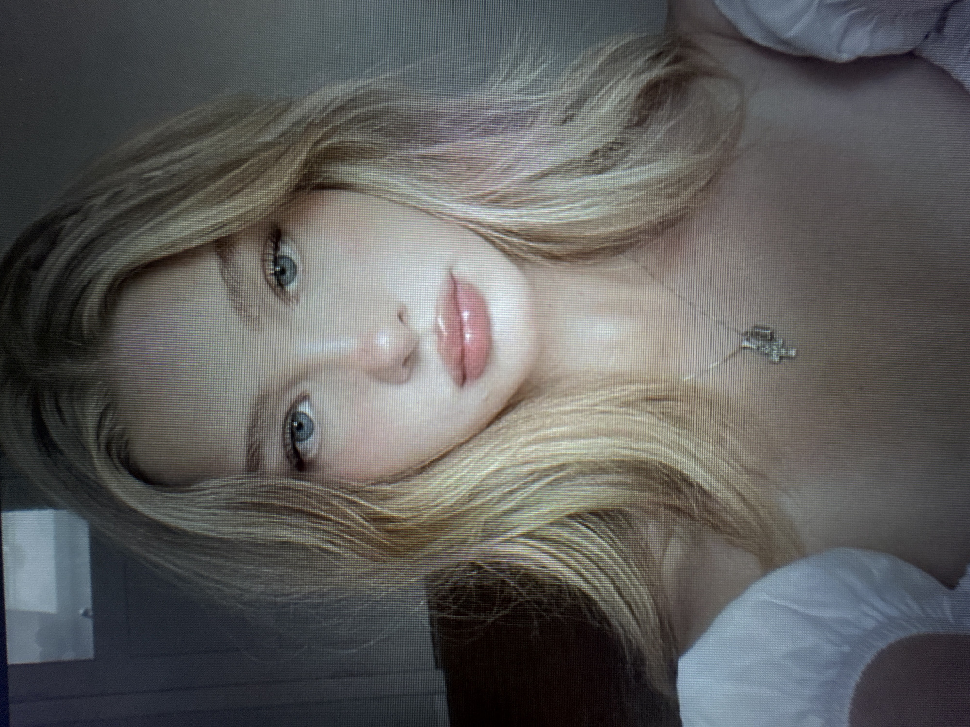 The media file presents a portrait of a young woman with long, flowing blonde hair, light blue or green eyes, and glossy, full lips. She looks directly at the viewer with a calm and serene expression. Her head is tilted, suggesting a relaxed pose, possibly while lying down. She wears a light-colored garment, which appears to be a top or dress with a ruffled or puffy shoulder/sleeve detail visible on the right side of the frame. A delicate silver-toned necklace with a small, intricate pendant rests on her chest.
The background is indistinct and muted, suggesting an indoor setting. The lighting is soft and diffused, highlighting her facial features.
A notable detail, evident in the main image and clearly emphasized by the provided close-up crops, is that the image itself is a photograph of a digital screen. This is indicated by the prominent pixel grid pattern visible throughout the picture, which impacts its overall sharpness and resolution. There is no visible text in the image.