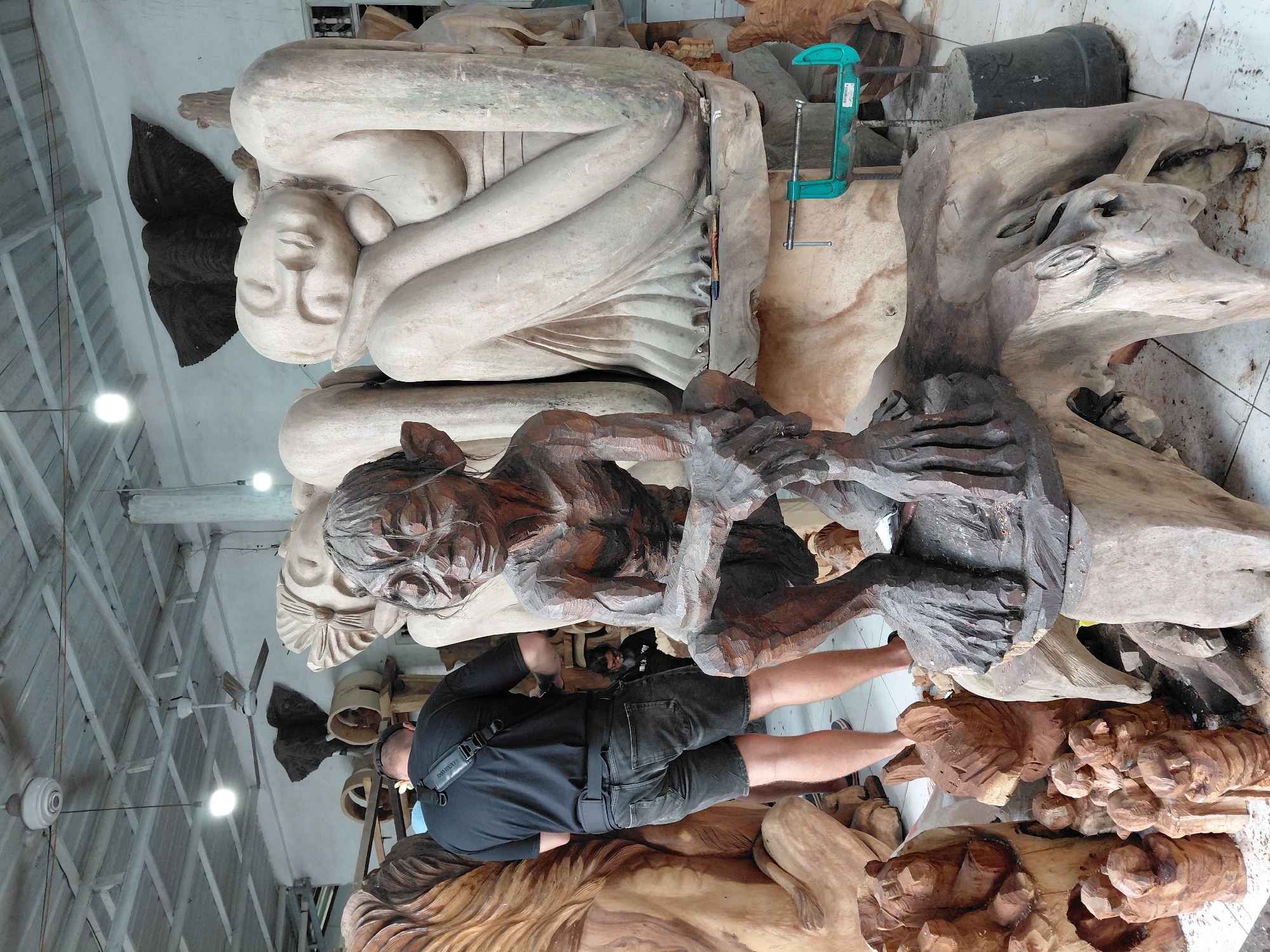 The image depicts a workshop or gallery filled with large wooden sculptures. In the foreground, a dark brown wooden sculpture of a creature with pointed ears, resembling Gollum from "The Lord of the Rings," is seated on a log. Behind it, a person wearing a black t-shirt and denim shorts stands with their back to the camera, facing other sculptures. To the right, a large, light-colored wooden sculpture of a reclining female figure with closed eyes is visible. In the background, more wooden carvings, including what appears to be a lion and several smaller figures, are arranged. A dark, elephant-shaped carving hangs on the wall. The environment appears to be an indoor space with a concrete floor, a corrugated metal ceiling, and visible lighting fixtures and fans. The overall impression is of an artisan's workshop, likely in Ubud District, Indonesia, given the prevalence of detailed wood carvings in that region.